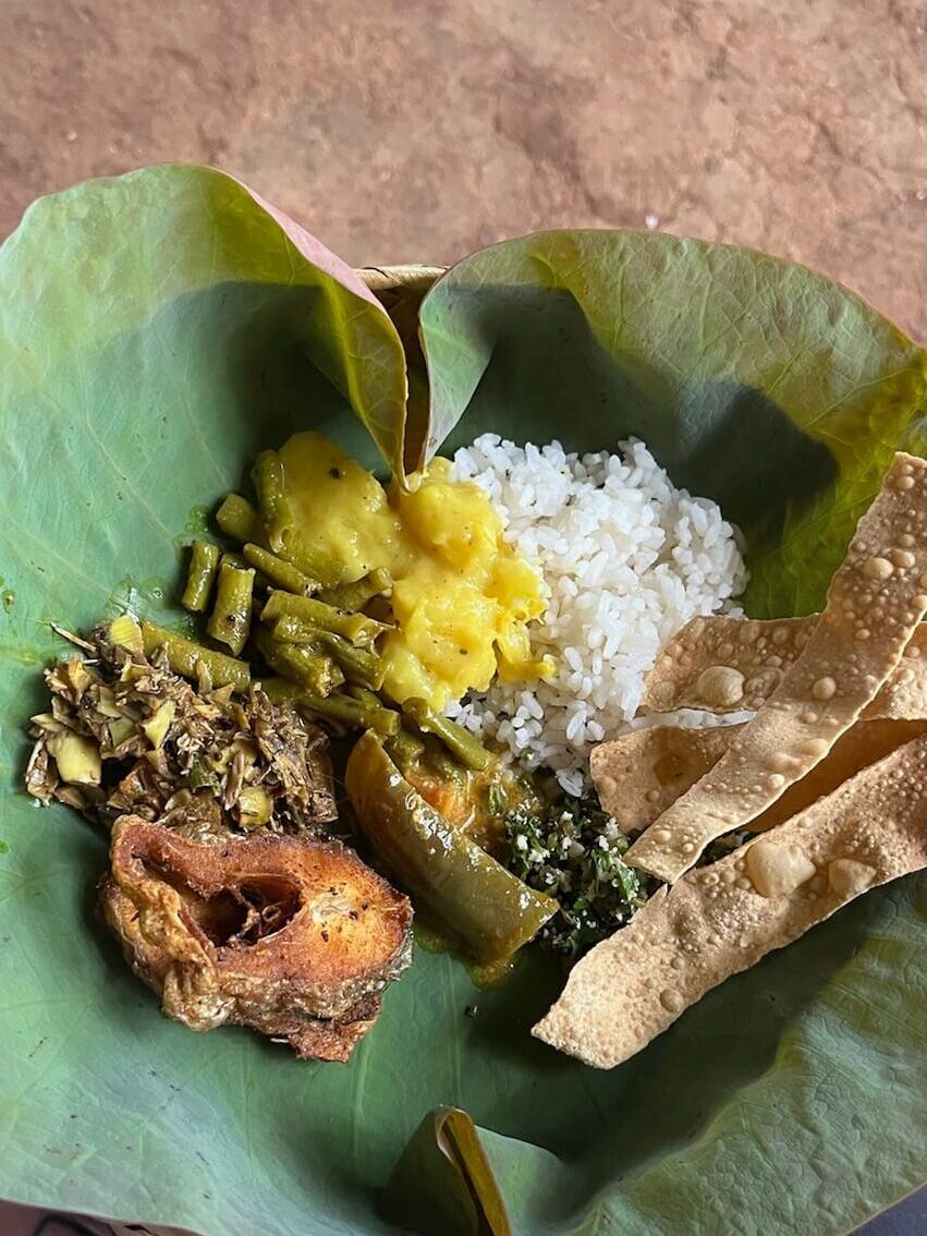 A large leaf used as a plate, covered in food: traditional flat bread, white rice, green bean curry, and various vegetables.