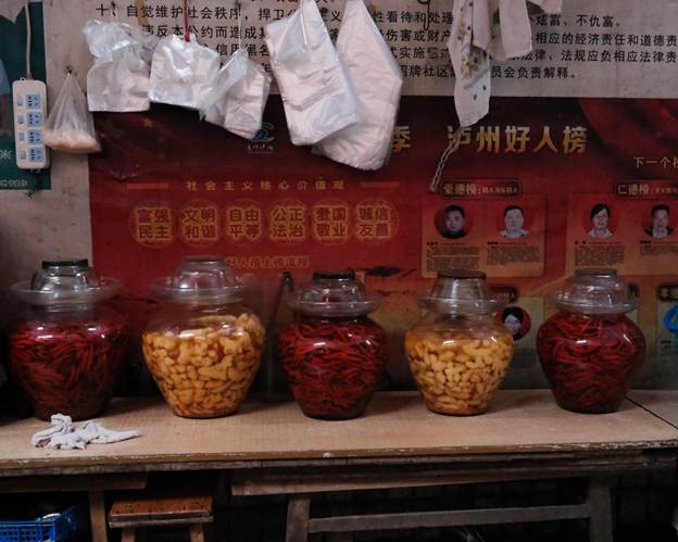 A makeshift wooden table adorned with 5 glass pickling jars filled with red Sichuan chilli peppers or yellow bamboo
