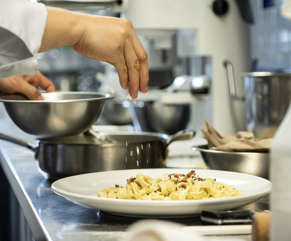 A hand dusting cheese on a pasta dish in a professional kitchen.