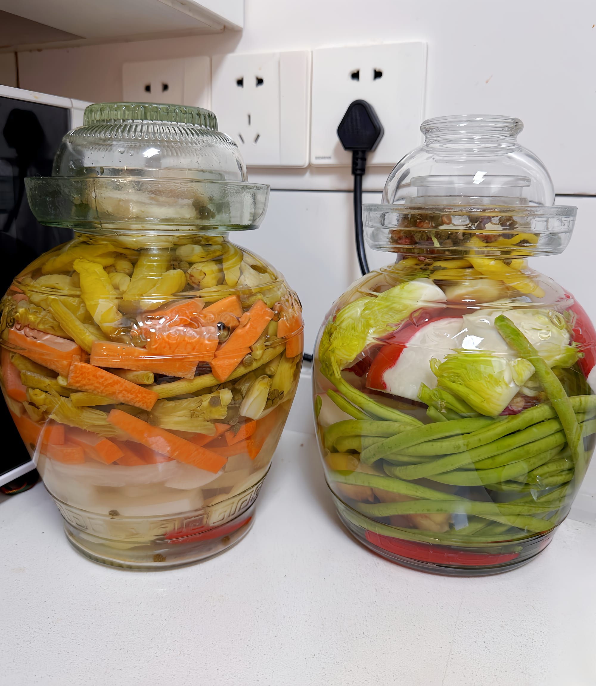 Two glass pickling jars full of carrots, long green beans, radishes, chili peppers, sitting on a white counter.