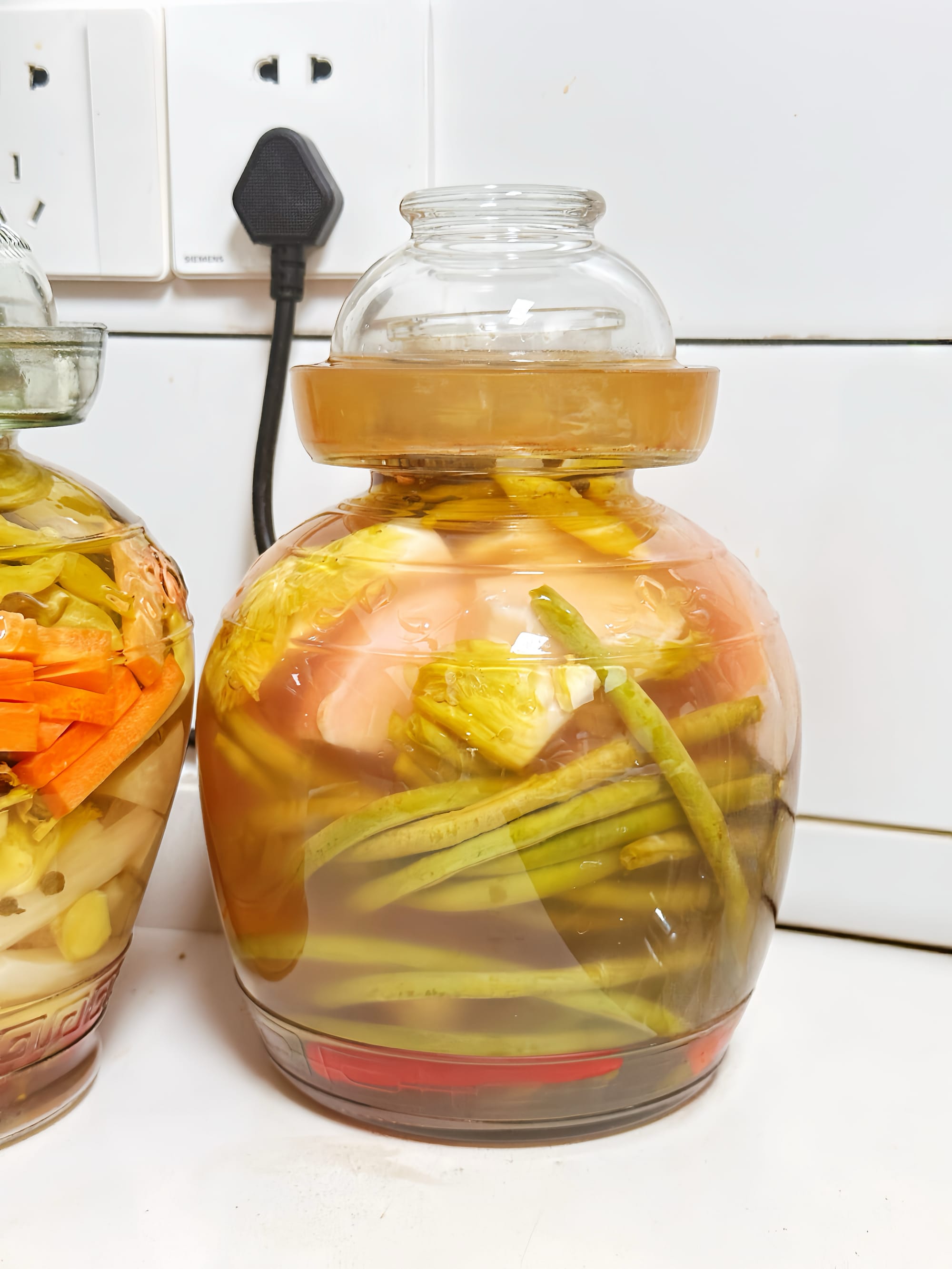 Two glass pickling jars at different stages, one to the left mostly clear towards the end of the process, and one in the centre of the frame turning milky.