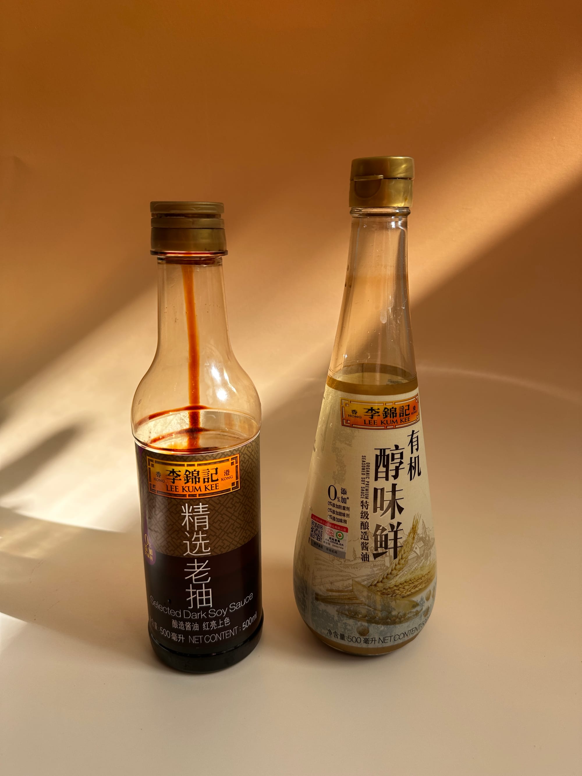 Two bottles of half-used soy sauce sit on a golden paper backdrop. One bottle shaped like a tear drop, the other like a short wine bottle. Brands are LEE KUM KEE