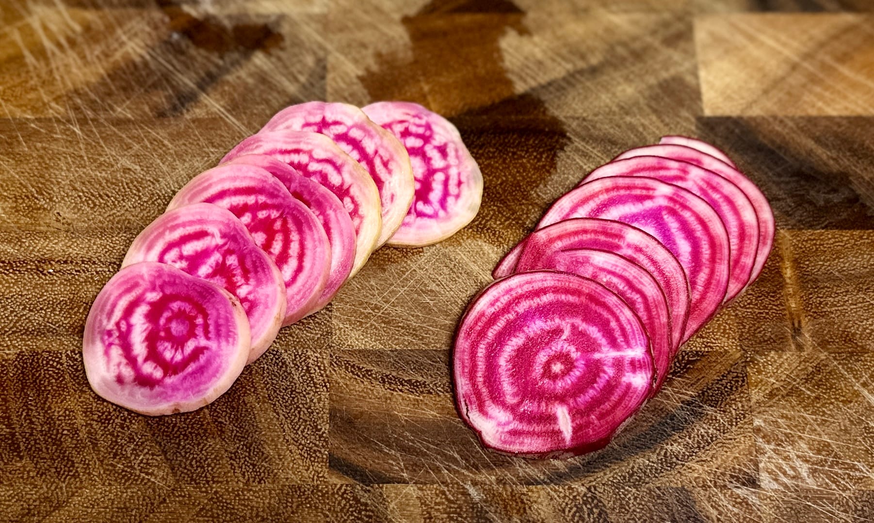 A brown wooden cutting board with two fanned out sets of sliced red beetroot. The beetroot slices are white with bright pink spiral and circular patterns.