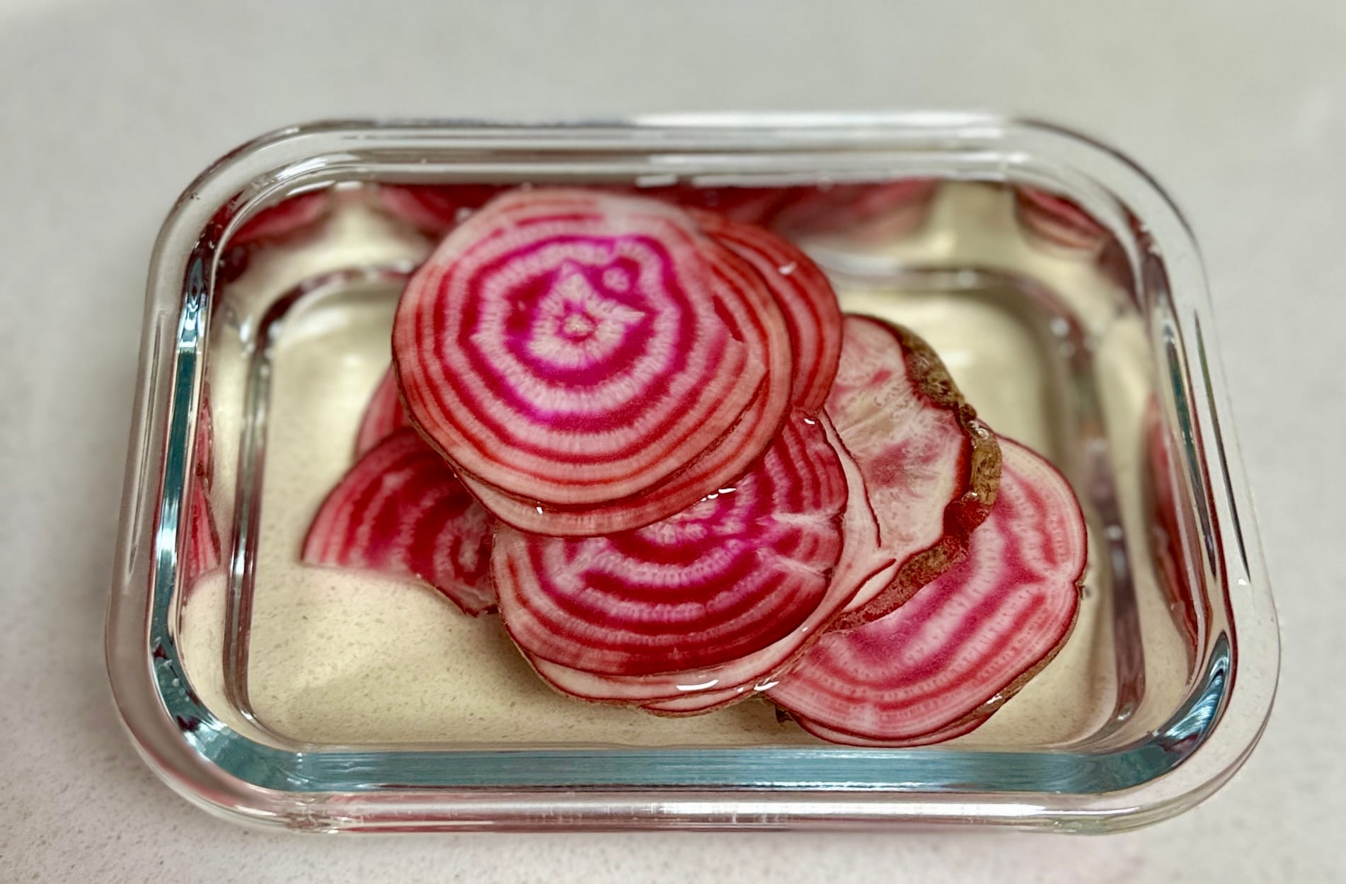 A clear glass container with vinegar and water with thinly sliced beetroot. Beetroot is white with pink spirals.