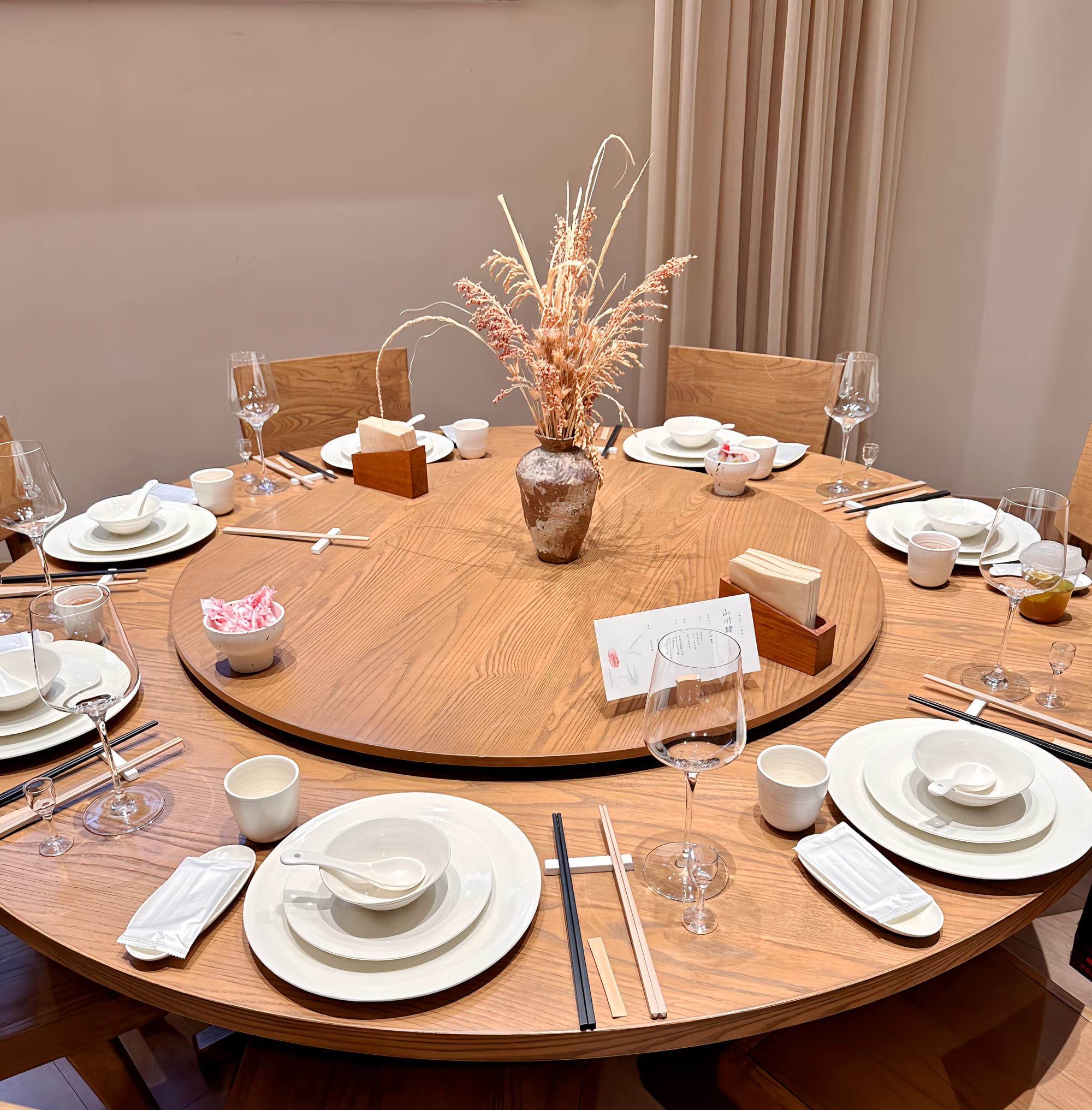 A traditional Chinese dinner table with lazy-susan in the middle surrounded by 7 complete seats with 2 plates, a bowl, soup spoon, cup, two pairs of chopsticks, wine glass, and hand towel. The table and lazy-susan are a light beige wood finish.