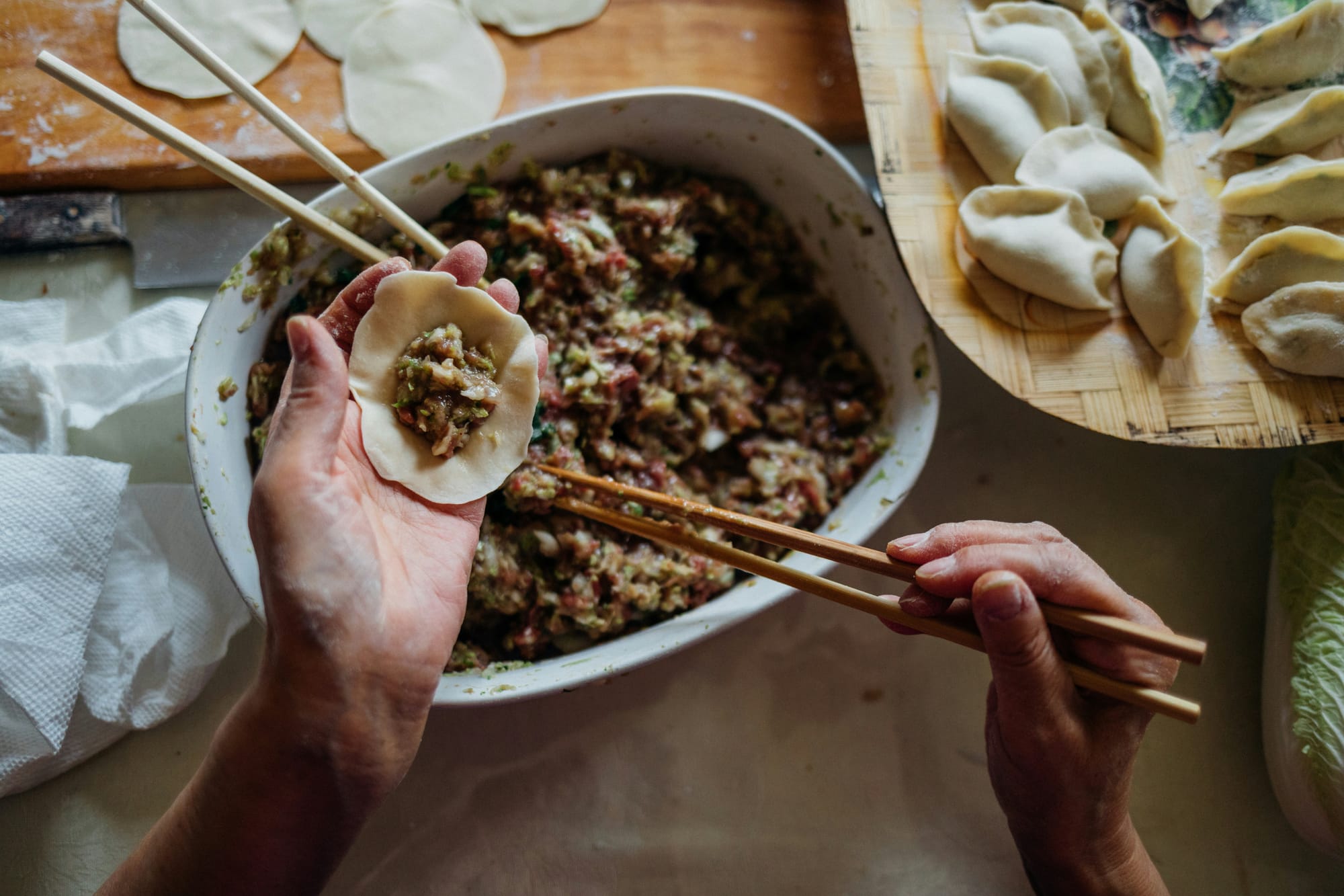 A kitchen counter, with a big casserole dish with minced pork and vegetables being used for dumpling filling. Two hands using chopsticks place filling into an open dumpling wrapper with previously made dumplings sitting on a tray next to the casserole dish.