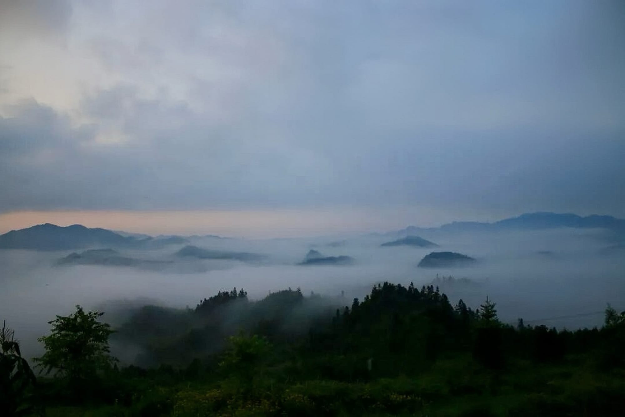 Forest covered mountains as far as the eye can see, enshrouded in mist with dark peaks cutting through under a gray and orange sky.