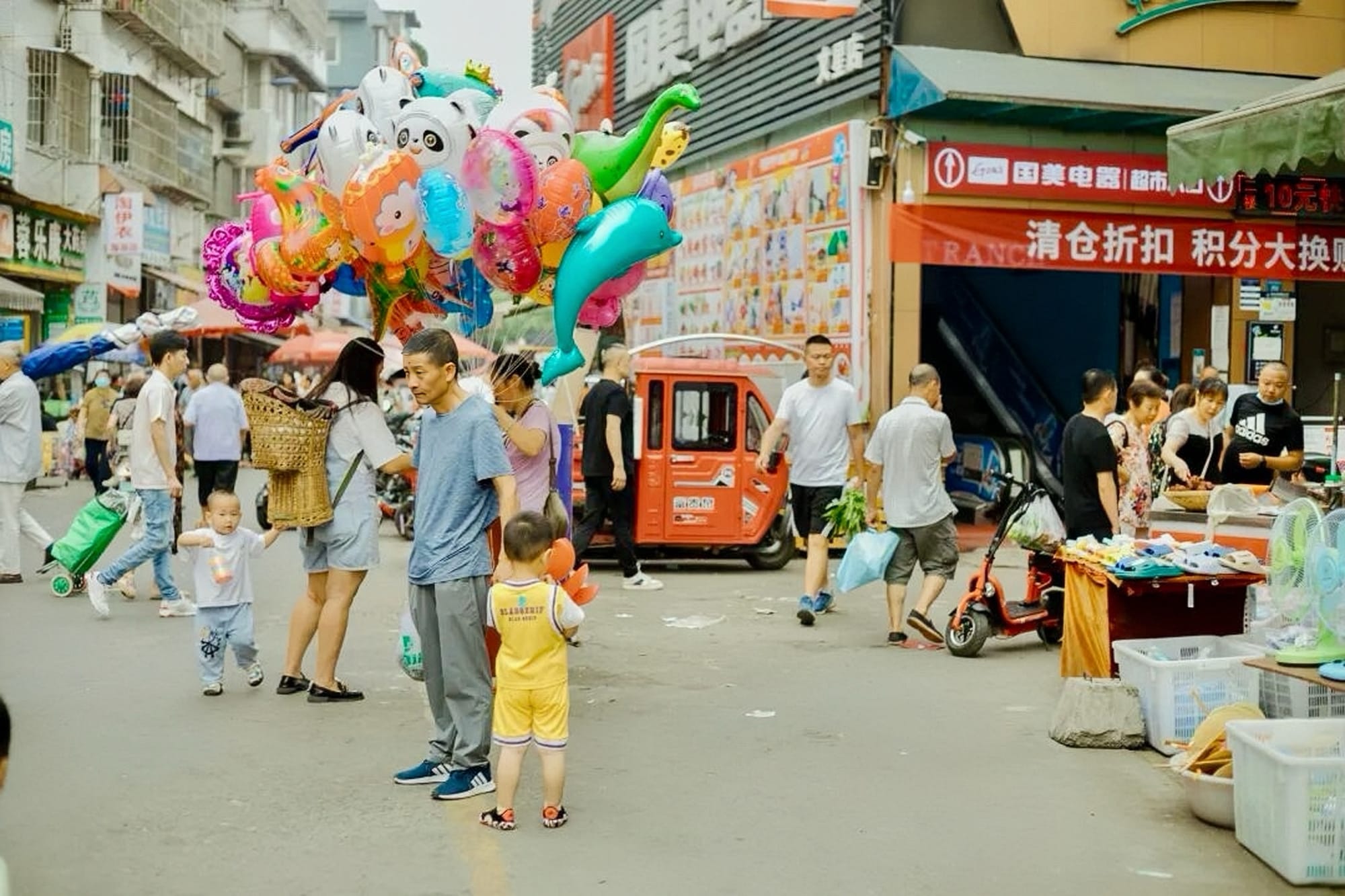 A chaotic market seen, people coming and going in every direction, a small stall to the right with a balloon vendor walking through the middle.