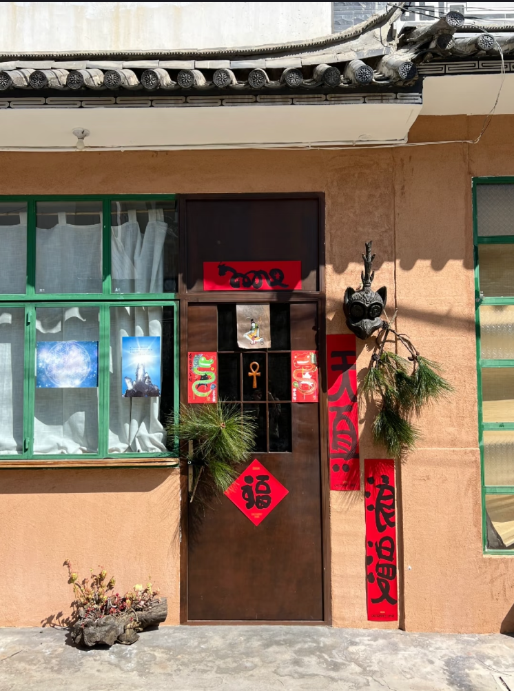 A red clay wall, with green-trimmed windows and a brown door centre frame. The door is flanked by red stickers with black Chinese writings. On the door and next to the door are also bows of pine needles.