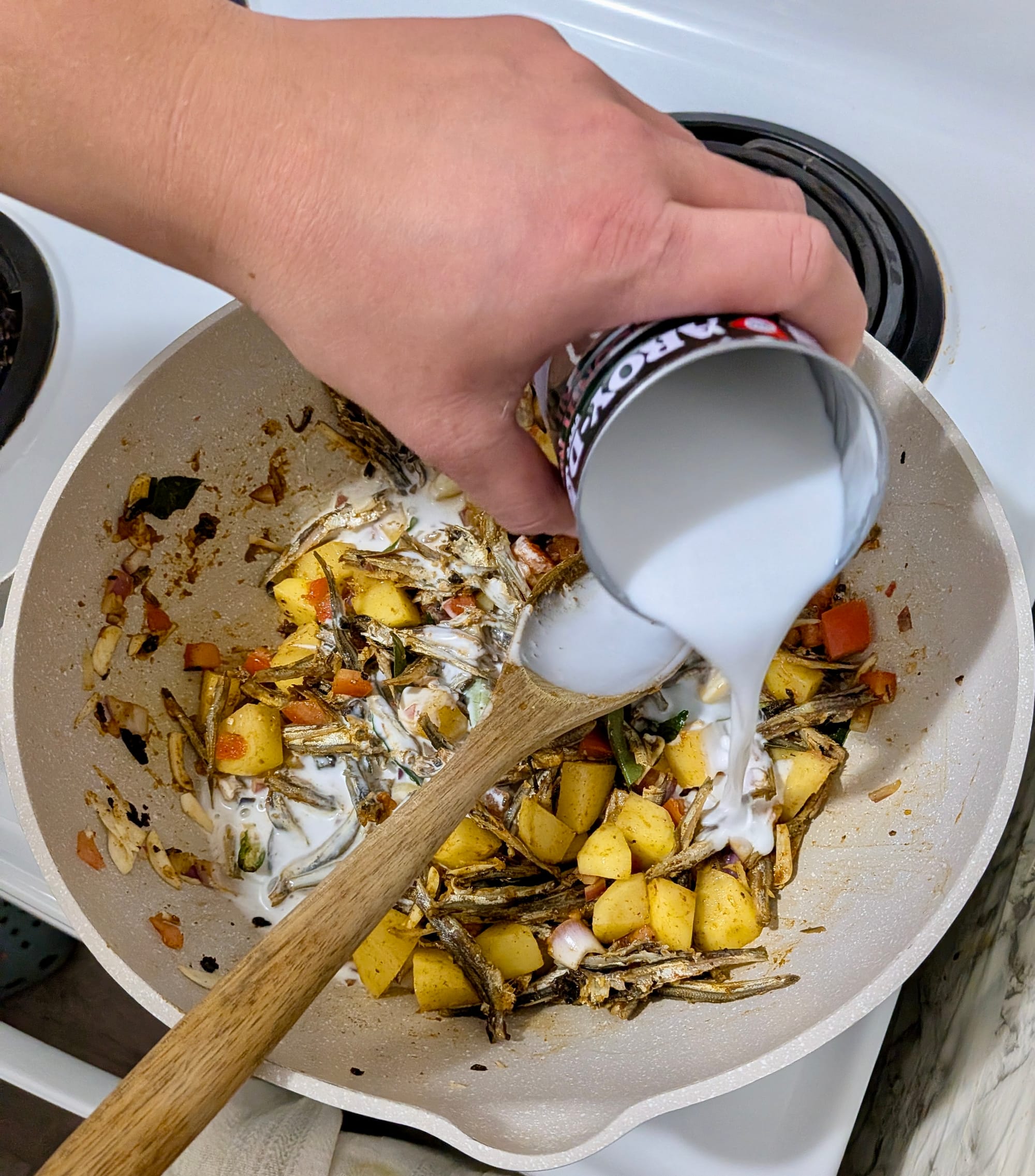 The ingredients for a breakfast curry in a pot on a white stove. A wooden spoon is used to mix the ingredients as coconut milk is added.