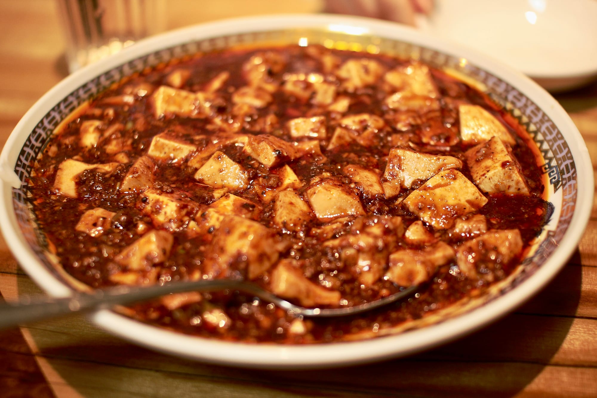 A small white dish decorated with a Chinese pattern on the rim, filled with mapo tofu (1cm cubed chunks of tofu covered in a deep red spicy sauce), with a metal spoon sticking out.