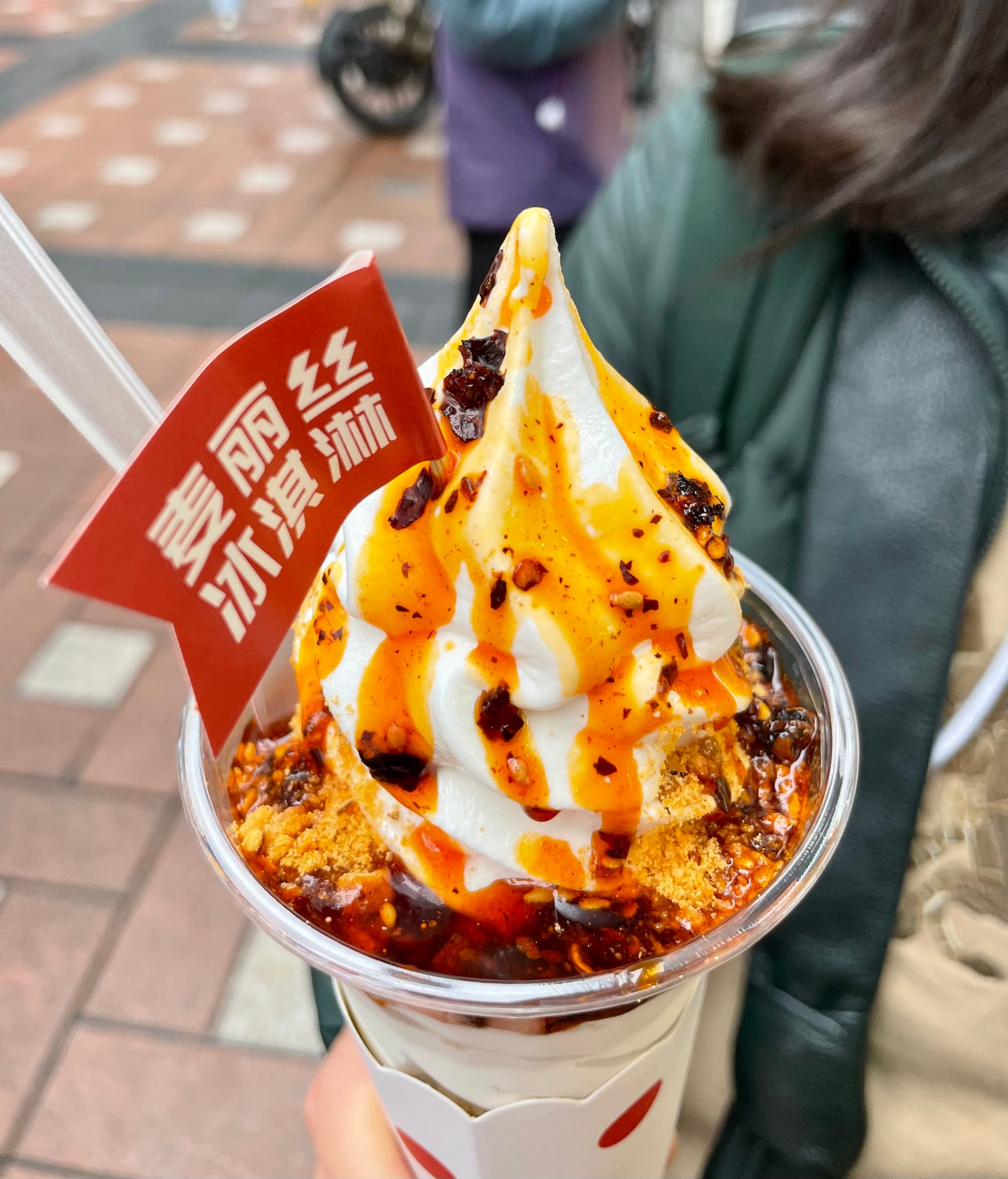 A white soft serve ice cream covered in mala spicy sauce with chili flakes and sesame. A small red flag pokes out the top with Chinese characters on it.