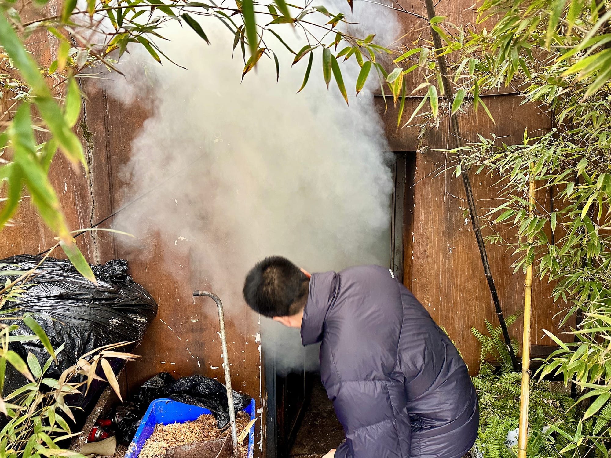 A brown building with a small door with smoke flooding out as Chef Tang stands before it peering in, ready to make adjustments to his culinary experiment inside.