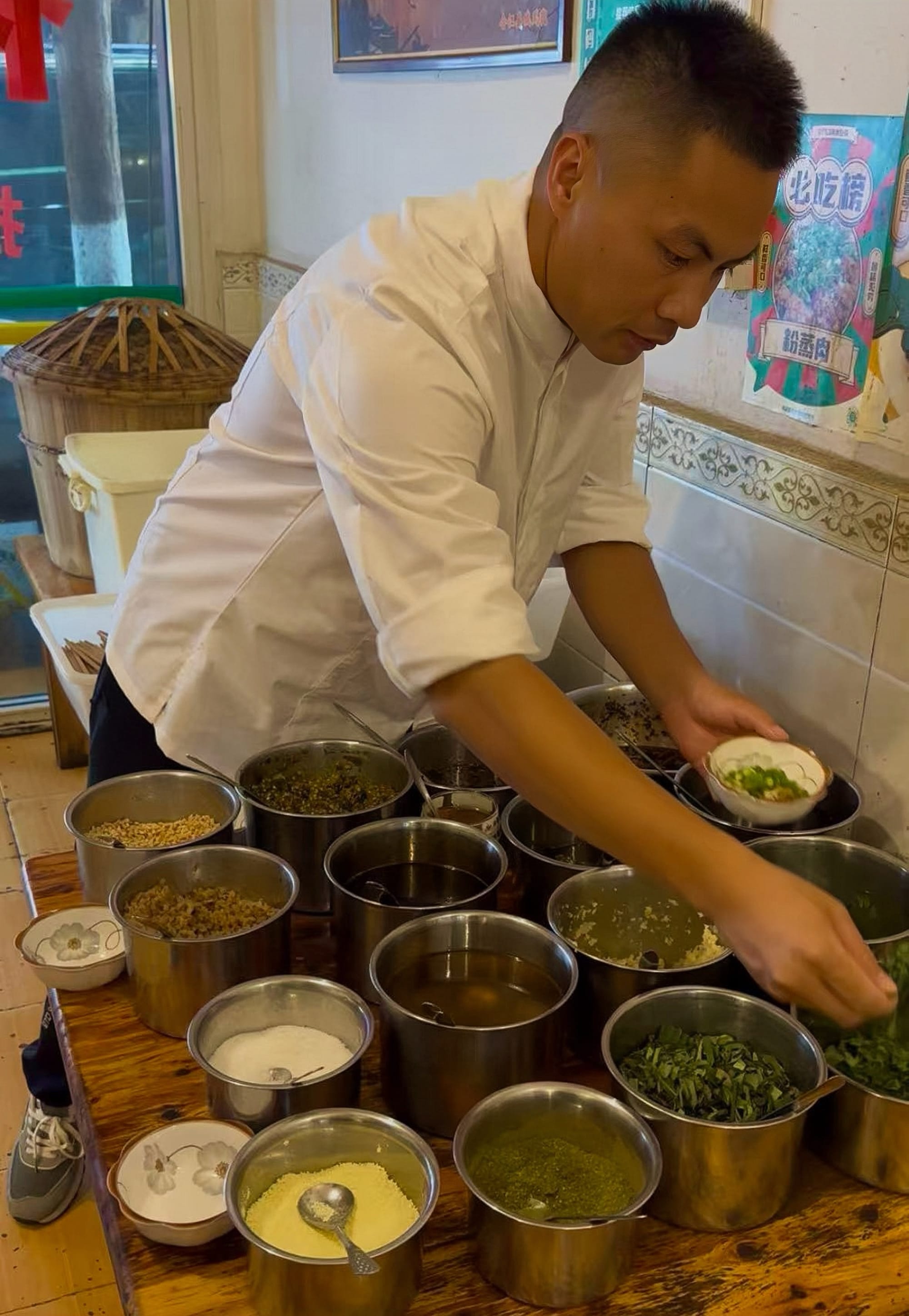 Chef Tang Bo leaning over a wooden table containing stainless steel cups, each filled with different spices, seasonings, and toppings.