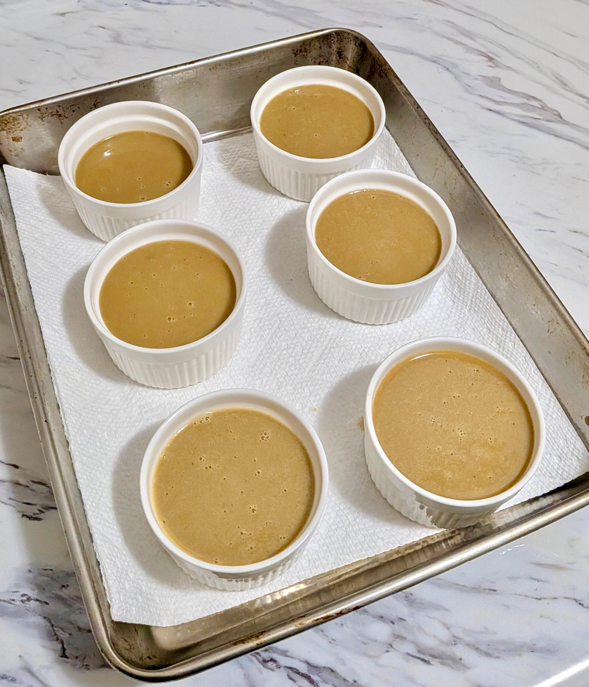 6 white ramekins rest on paper towel within a deep cooking sheet. The ramekins are filled with Watalappam mixture.