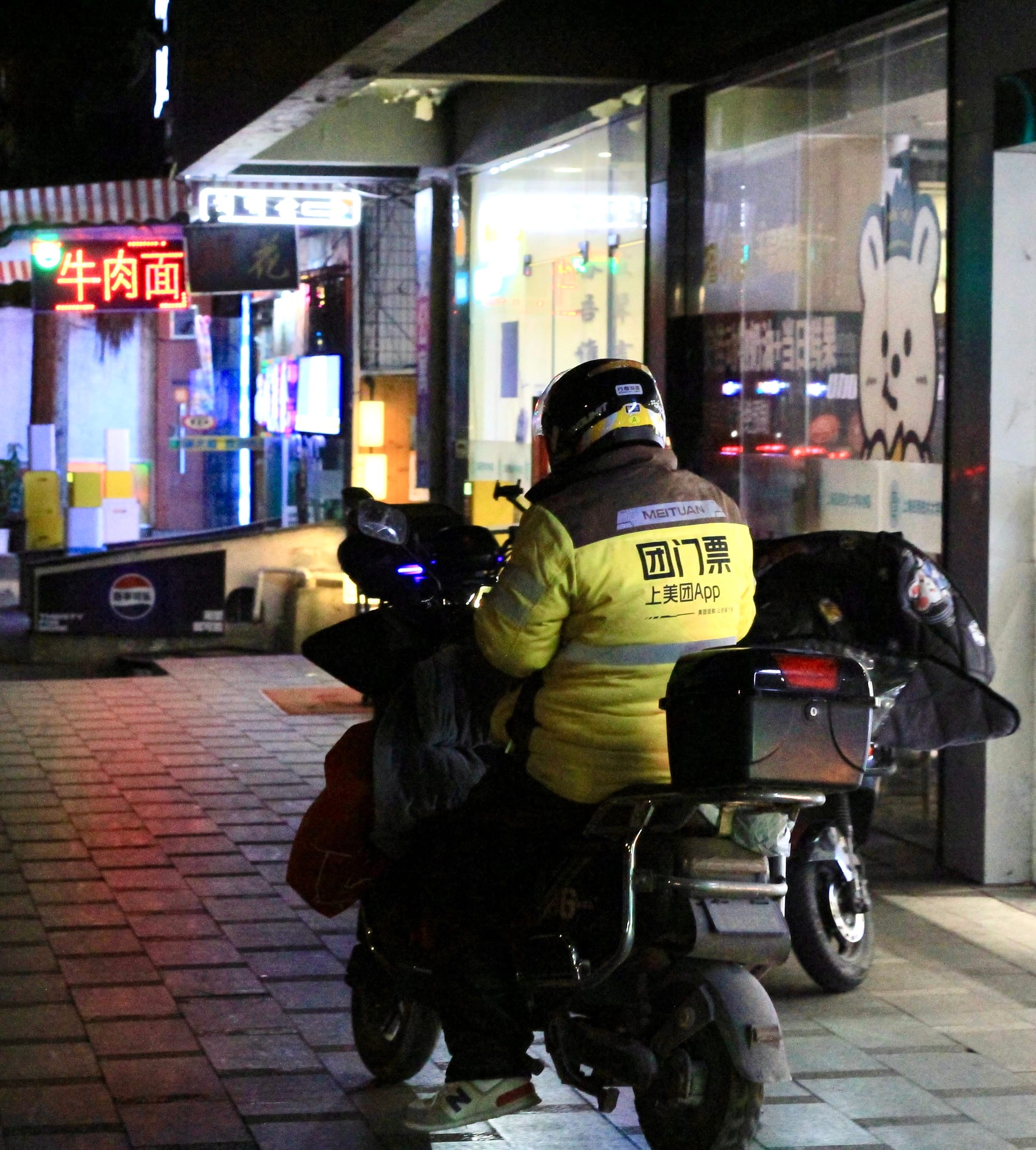 Night time, a MeiTuan delivery driver in their yellow winter coat sits on a scooter, browsing his phone on a Shanghai sidewalk next to stores.