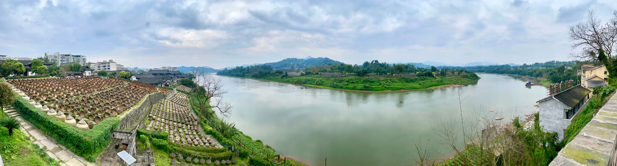 The Chishui river bends before a bank of soy sauce urns by the hundred on an old green hill. Across the river are green banks and misty foothills.