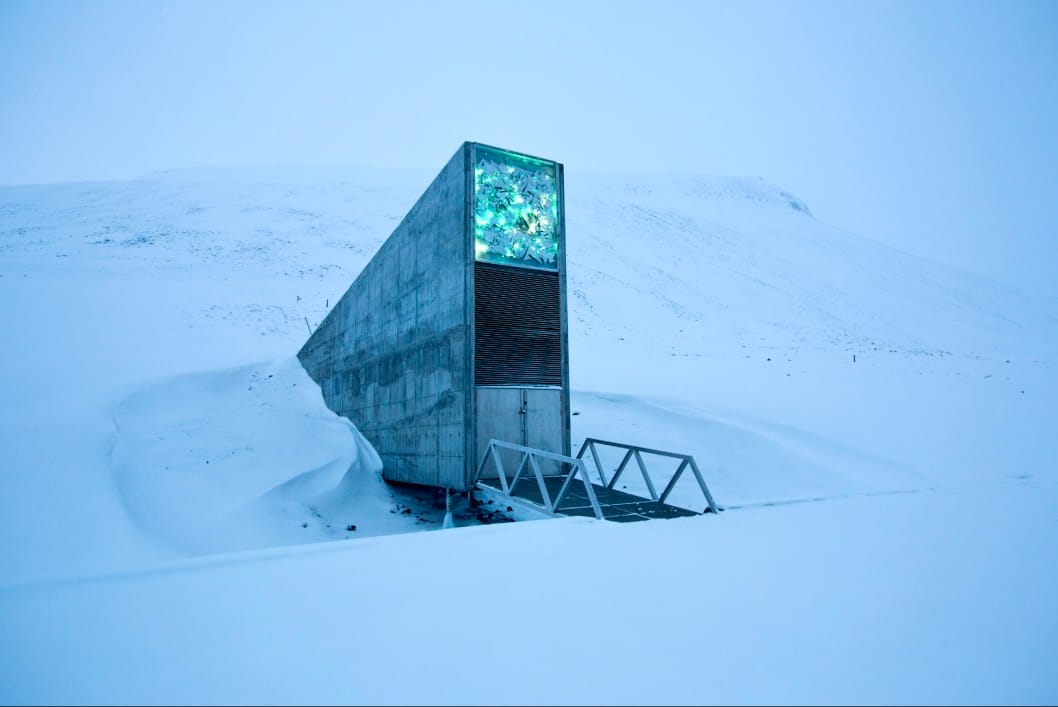 The concrete entrance to the Svalbard Global Seed Vault pokes out of a snowy mountain backdrop. A small metal footbridge leads to two metal doors topped by a giant industrial intake fan and a colorful sign of a mosaic of silvery blue and green.