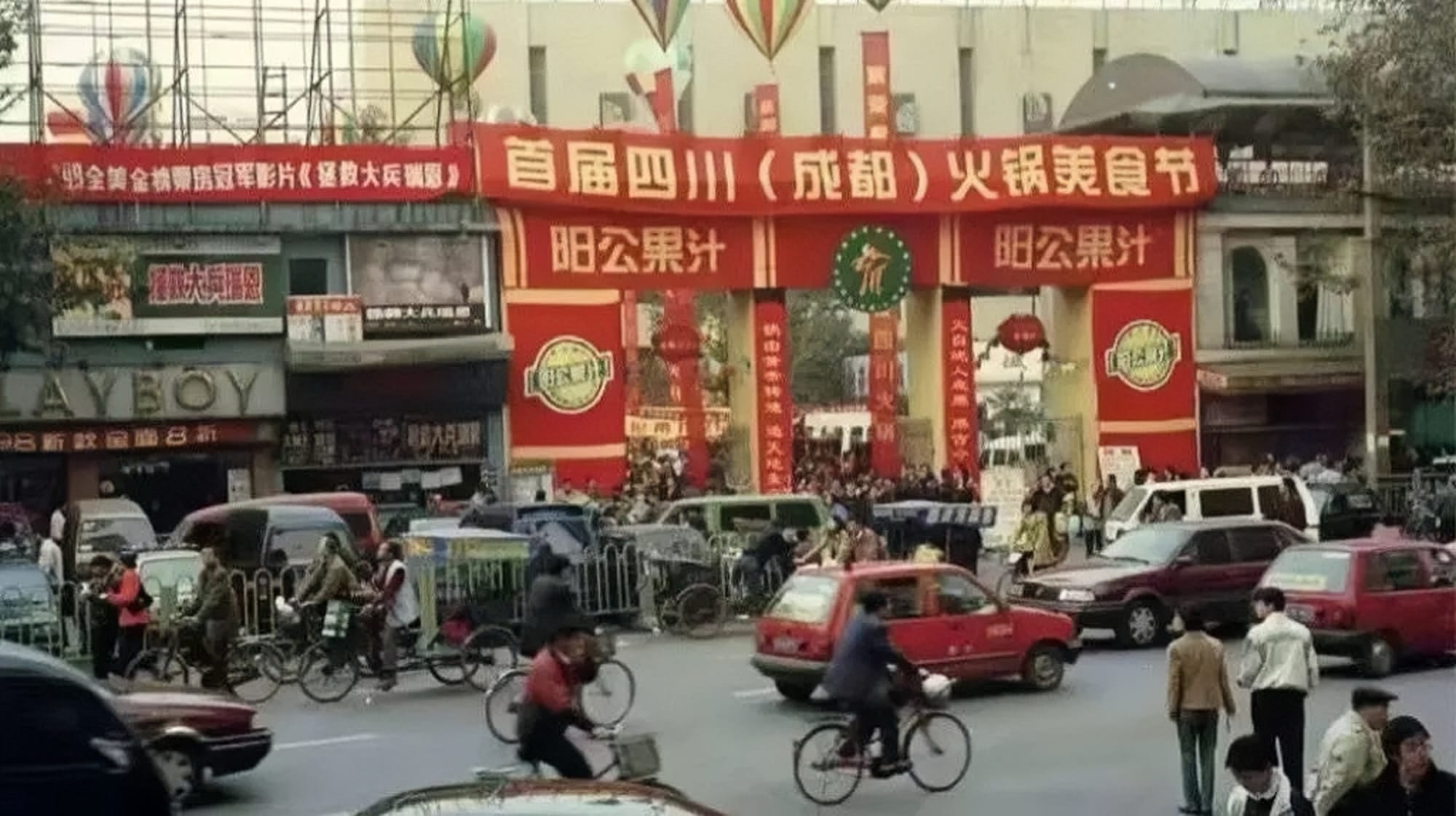 A busy street in Chengdu, filled with pedestrians, bicycles, and cars. Across the road are small shops, with a large gate in the middle. The gate is clad with big red banners covered in golden writing in Chinese characters, welcoming people to the first Chengdu Hotpot festival.