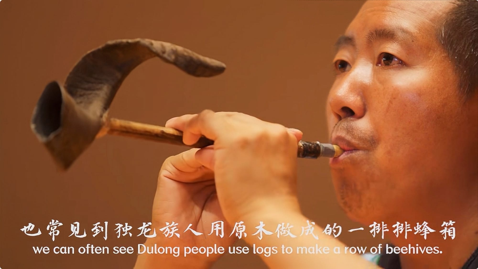 A screen capture of a Dulong man blowing into a traditional instrument made from beehives.