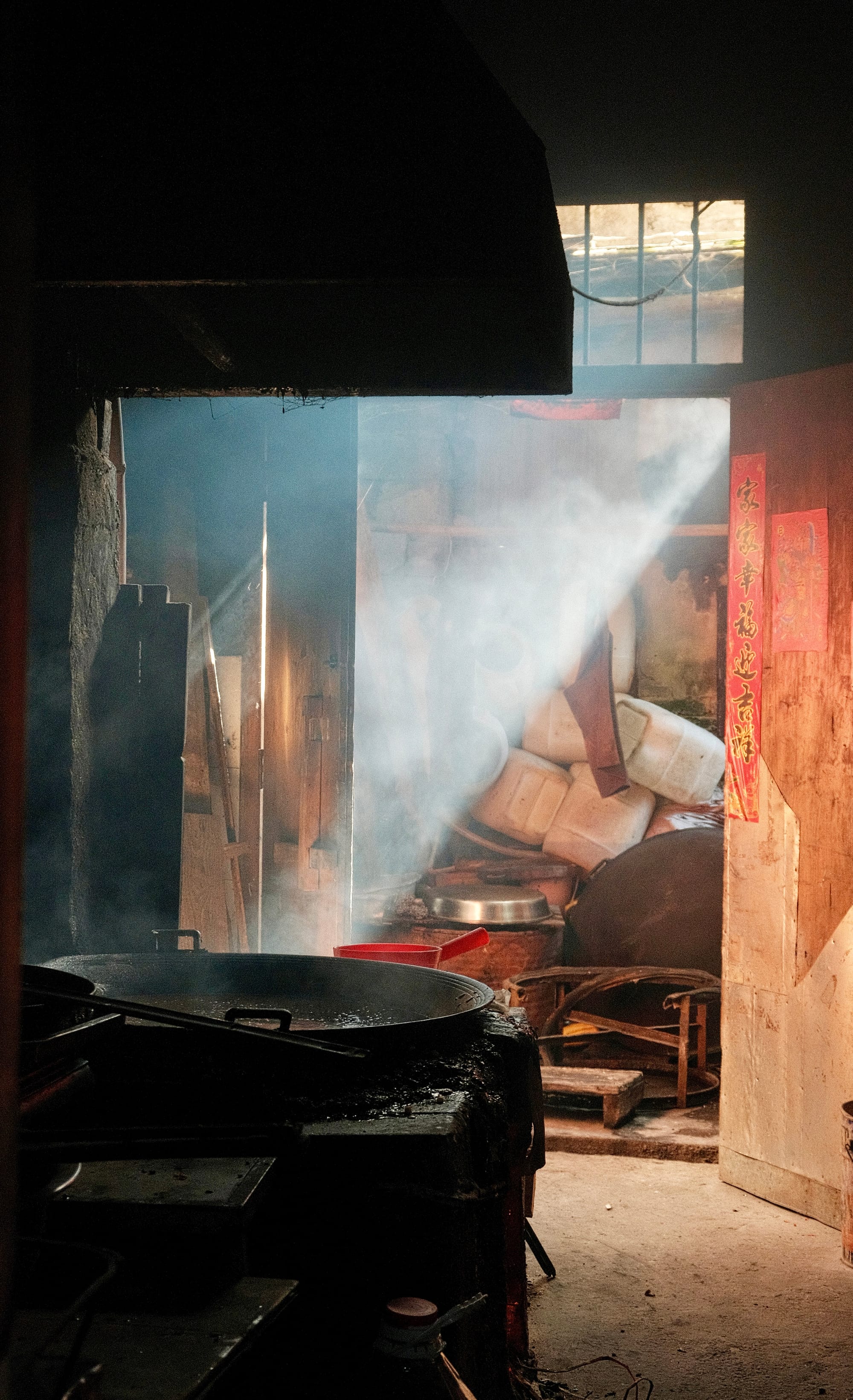 A small pine blossom candy shop called Rongdi in Yangshuo - a traditional brick stove with a large iron inlaid wok sits steaming, with a messy wooden closet in the background as sunlight pours through the window