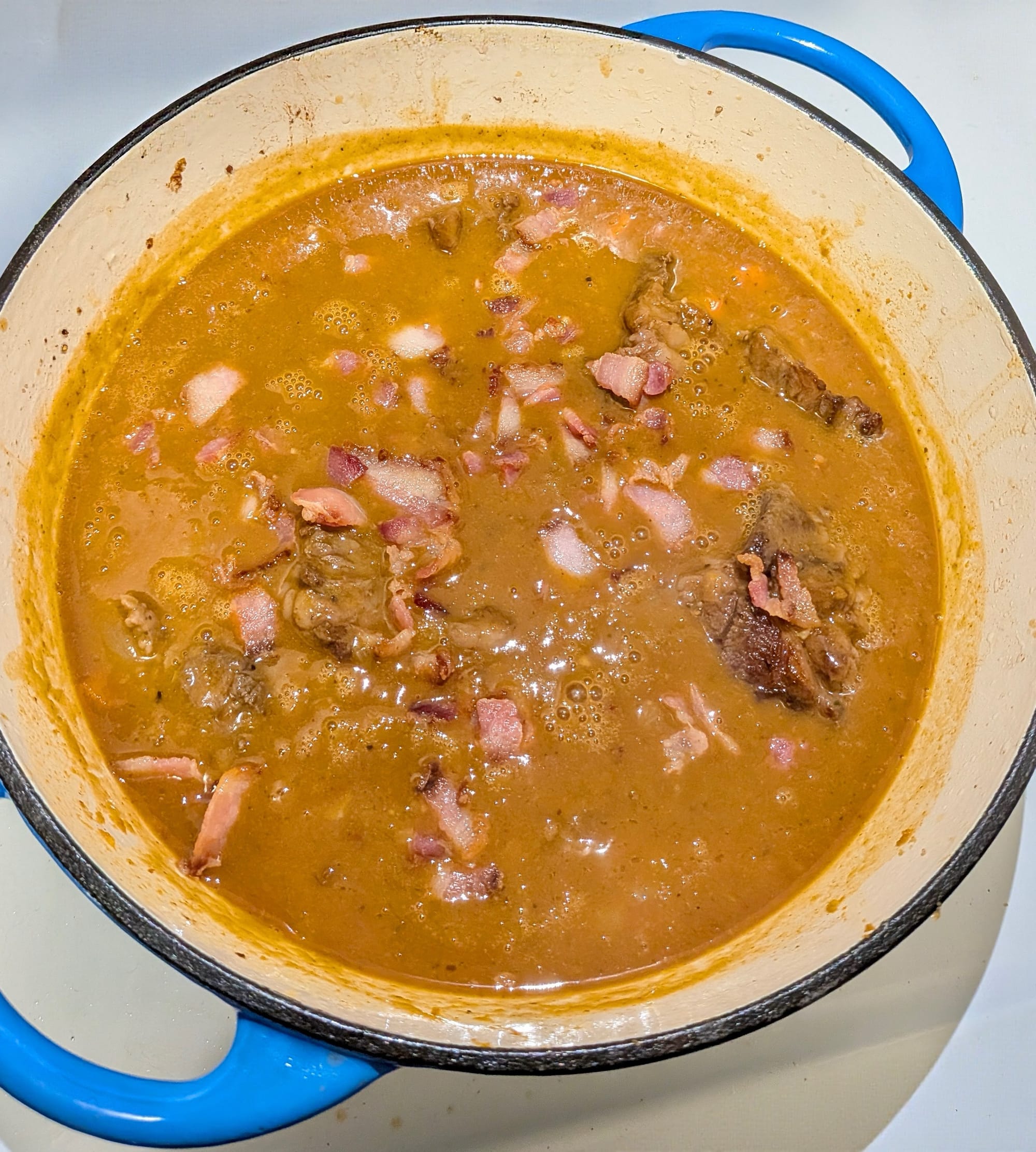 Stoofvlees simmering in a pot - a thick orange-brown gravy with chunked beef, bacon, and potato.