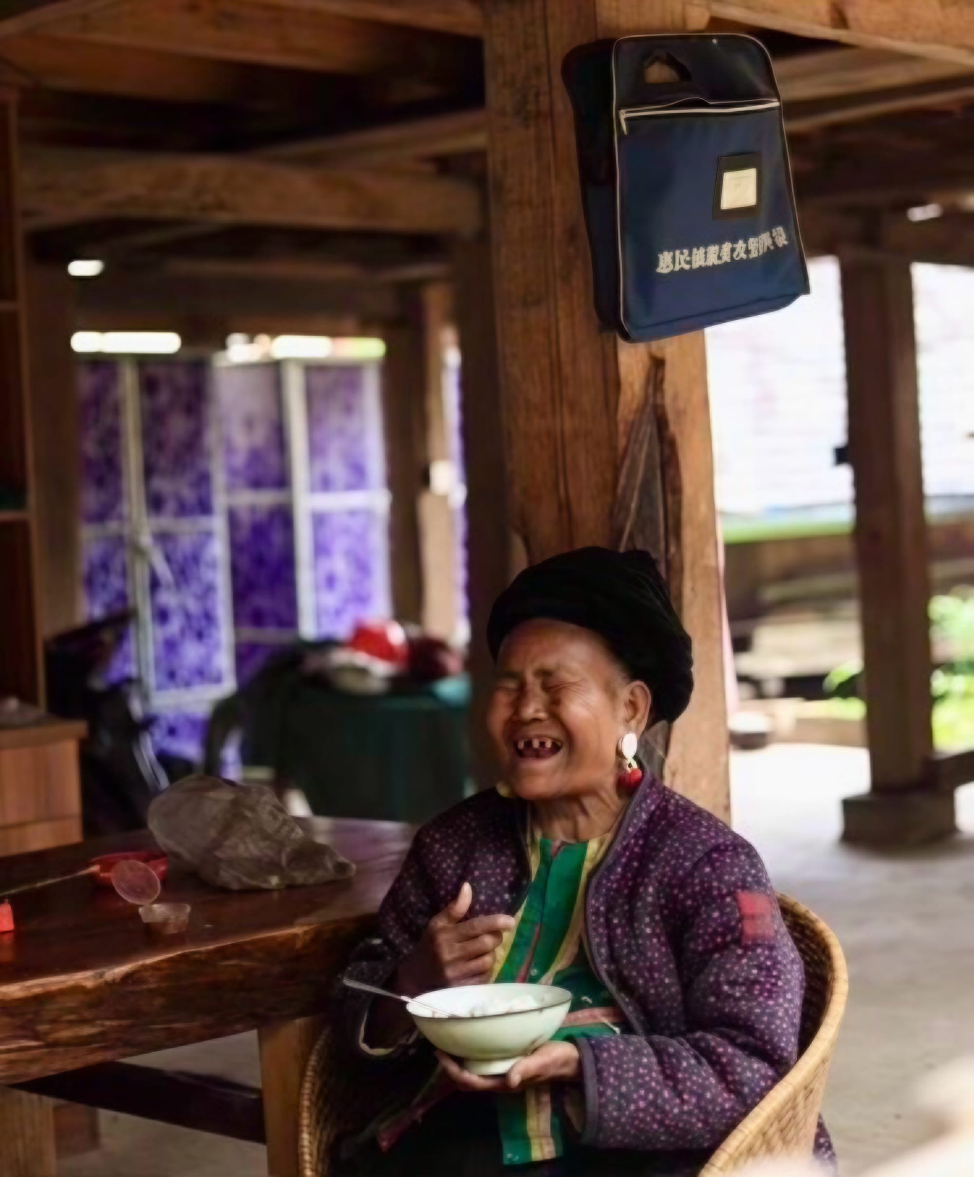 An elderly Blang woman sitting in a bamboo chair eating from a white bowl, laughing.