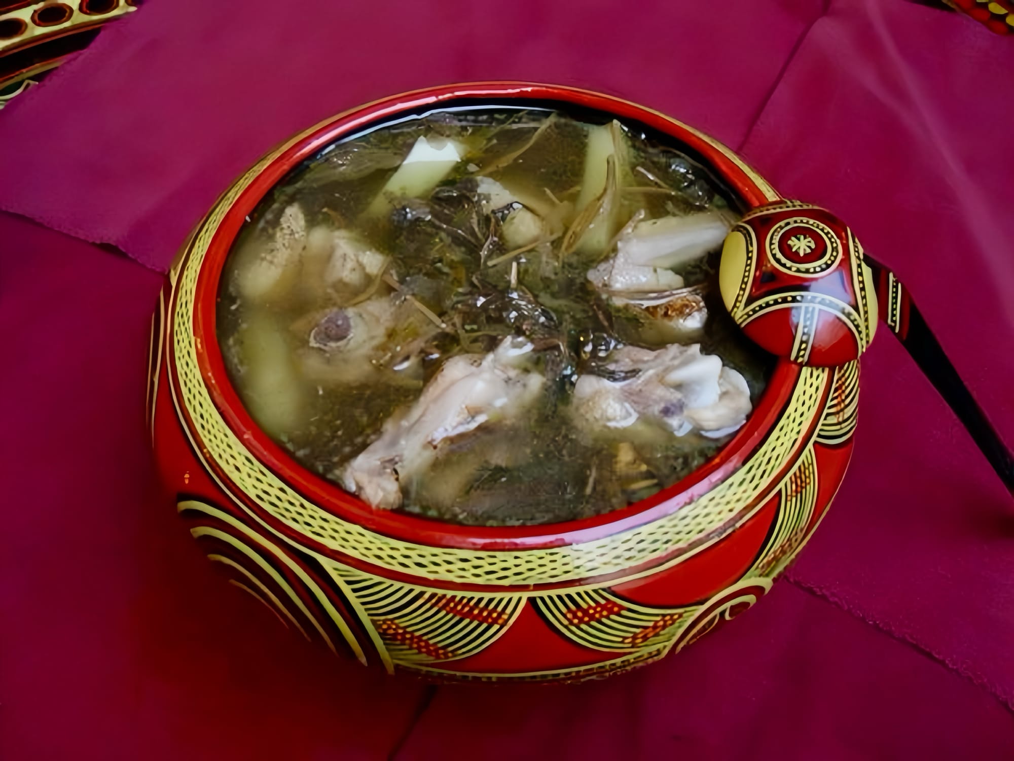 An intricately designed red with yellow pattern design soup bowl with matching spoon, filled with soup.