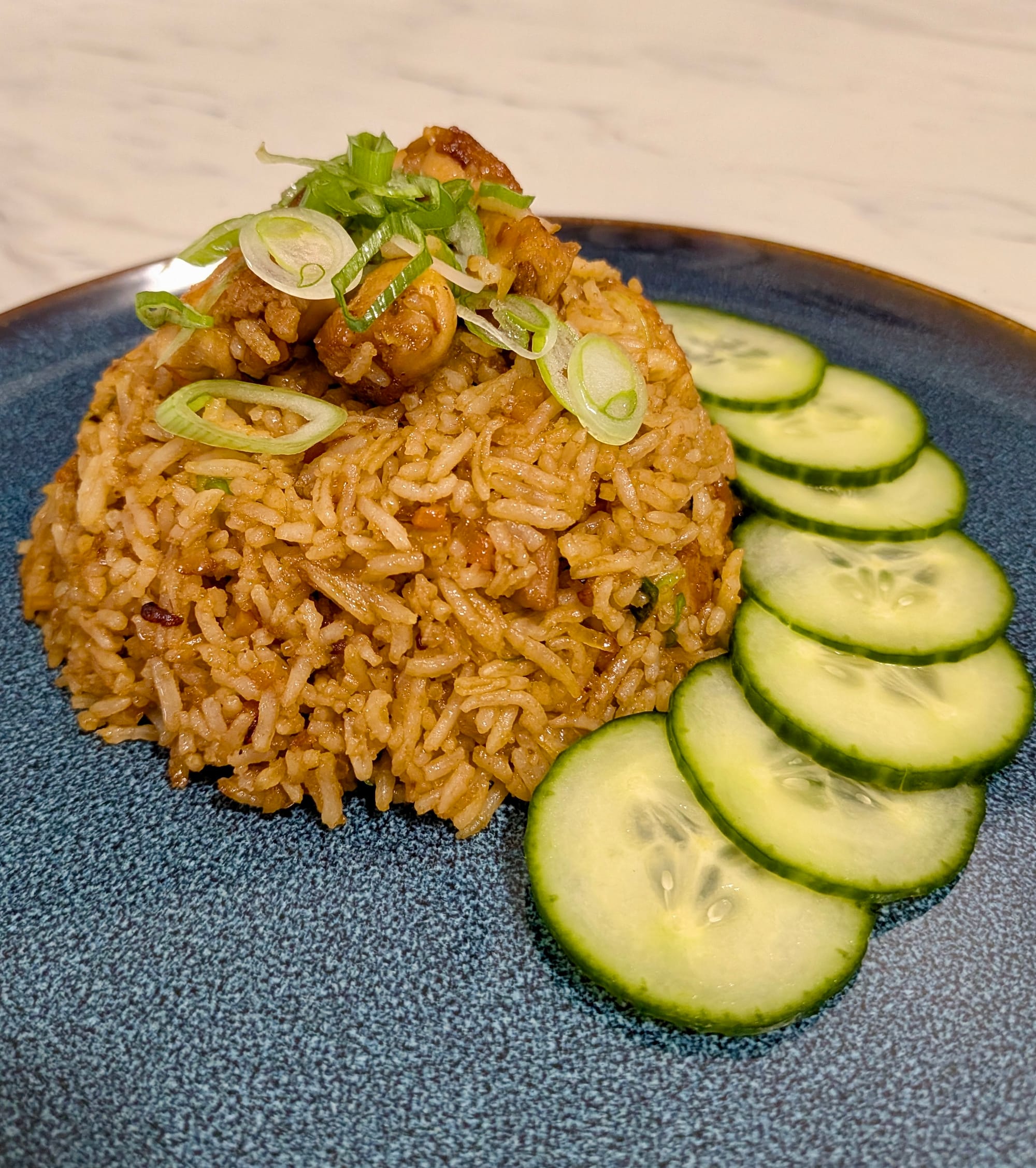 Nasi Goreng fried rice with diced spring onion sprinkled on top and diced cucumbers around the side on a blue plate.