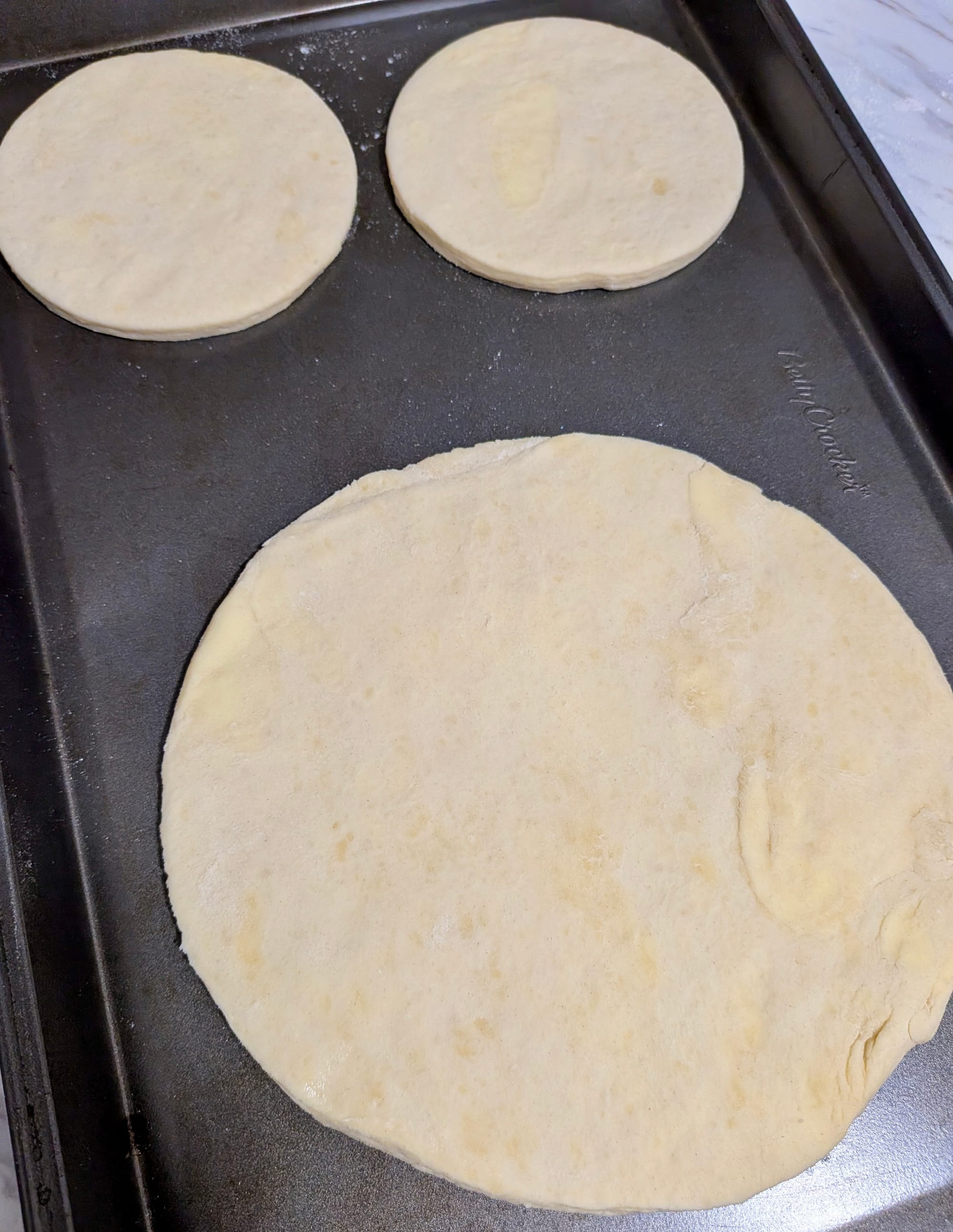 Three light colored dough circles on a pan, two are smaller around 5cm across, the other is larger around 15cm across.