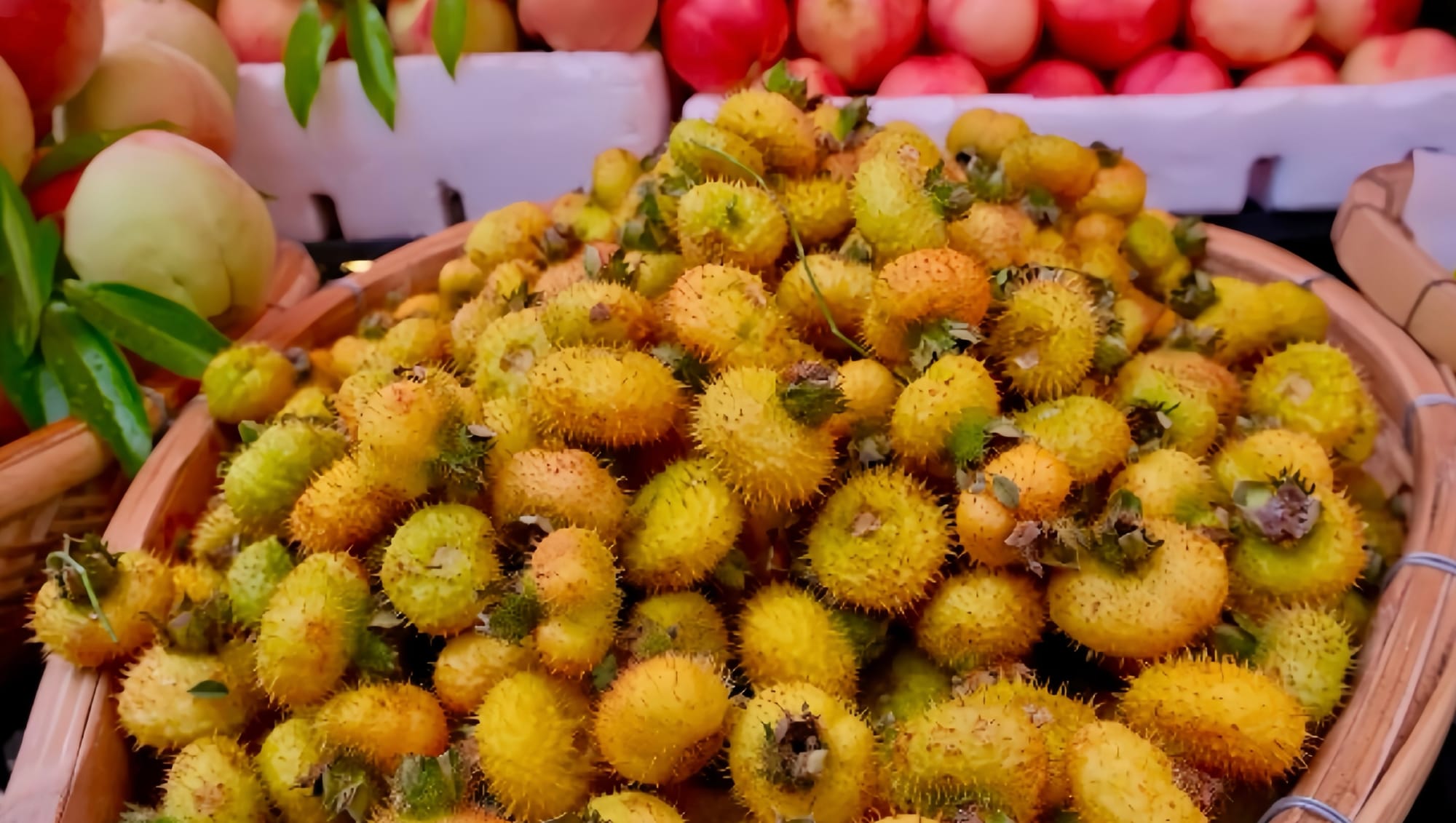 A basket of fresh yellow-greenish spiky cili fruits next to baskets of white peaches.