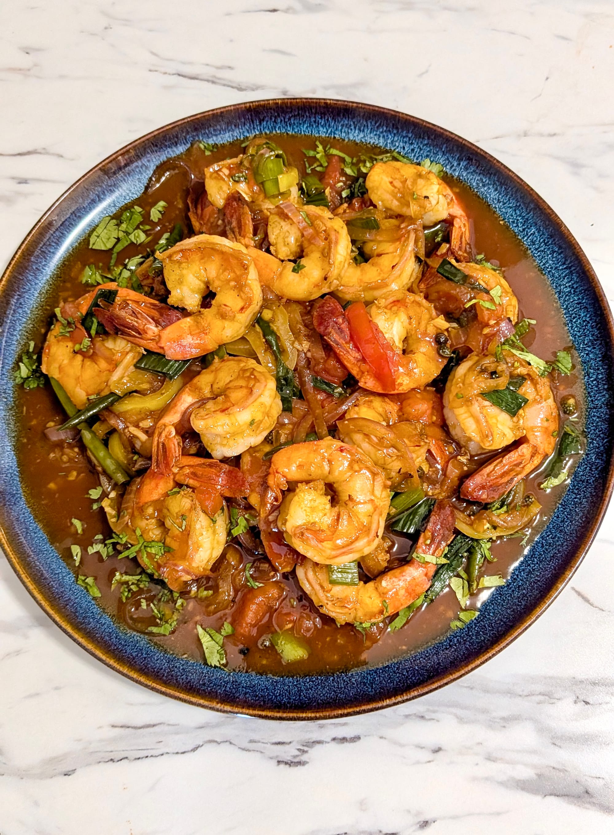 Directly over top view of Mala Devilled Prawns, golden-red prawns with mixed chopped vegetables and a brown sauce on a blue plate.