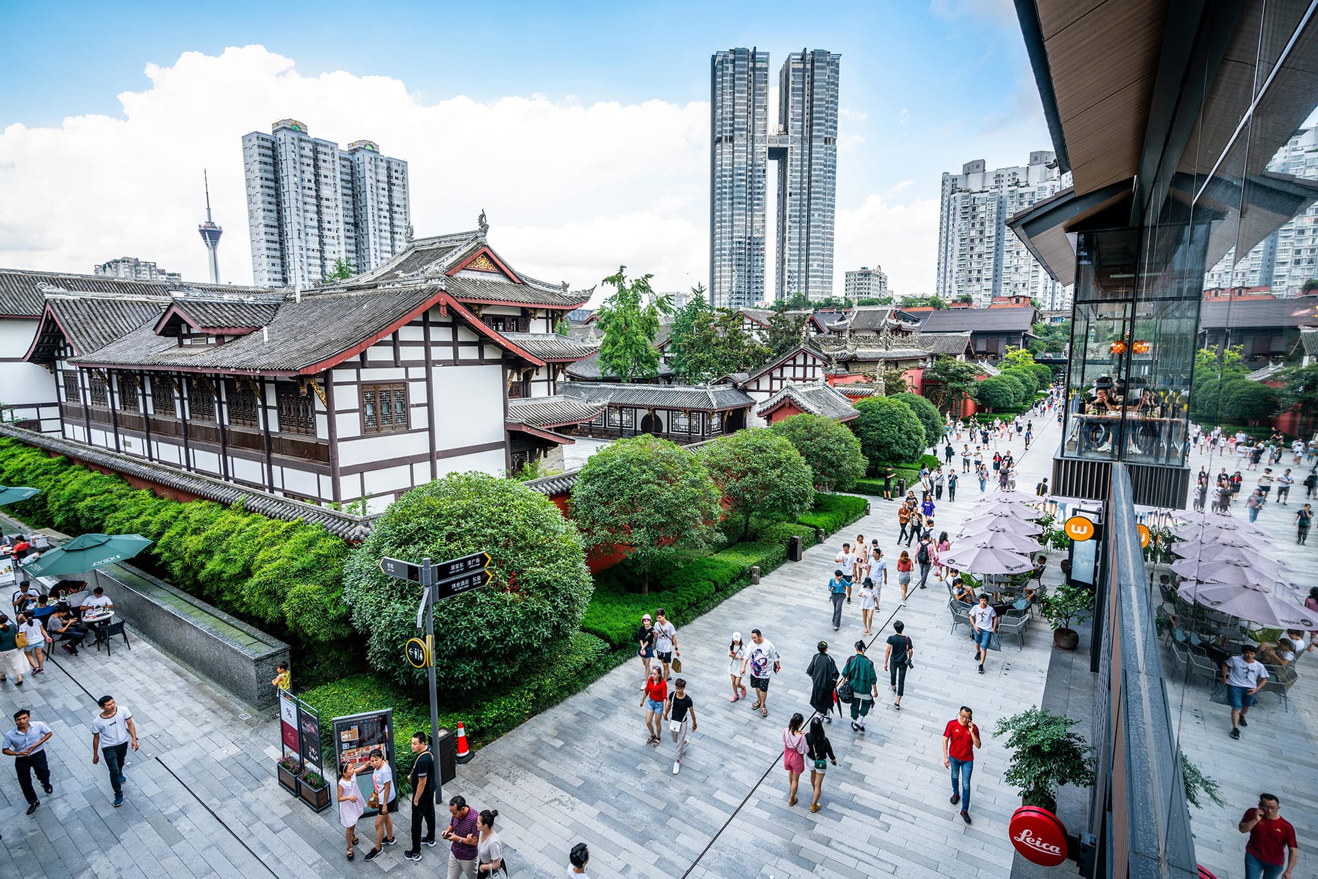 A view of TaiKooLi in Chengdu from a second-floor terrace. Ancient-style buildings flanked by trees and shrubs and stone walkways full of people. In the distance, silver sky-scraper buildings line the sky.
