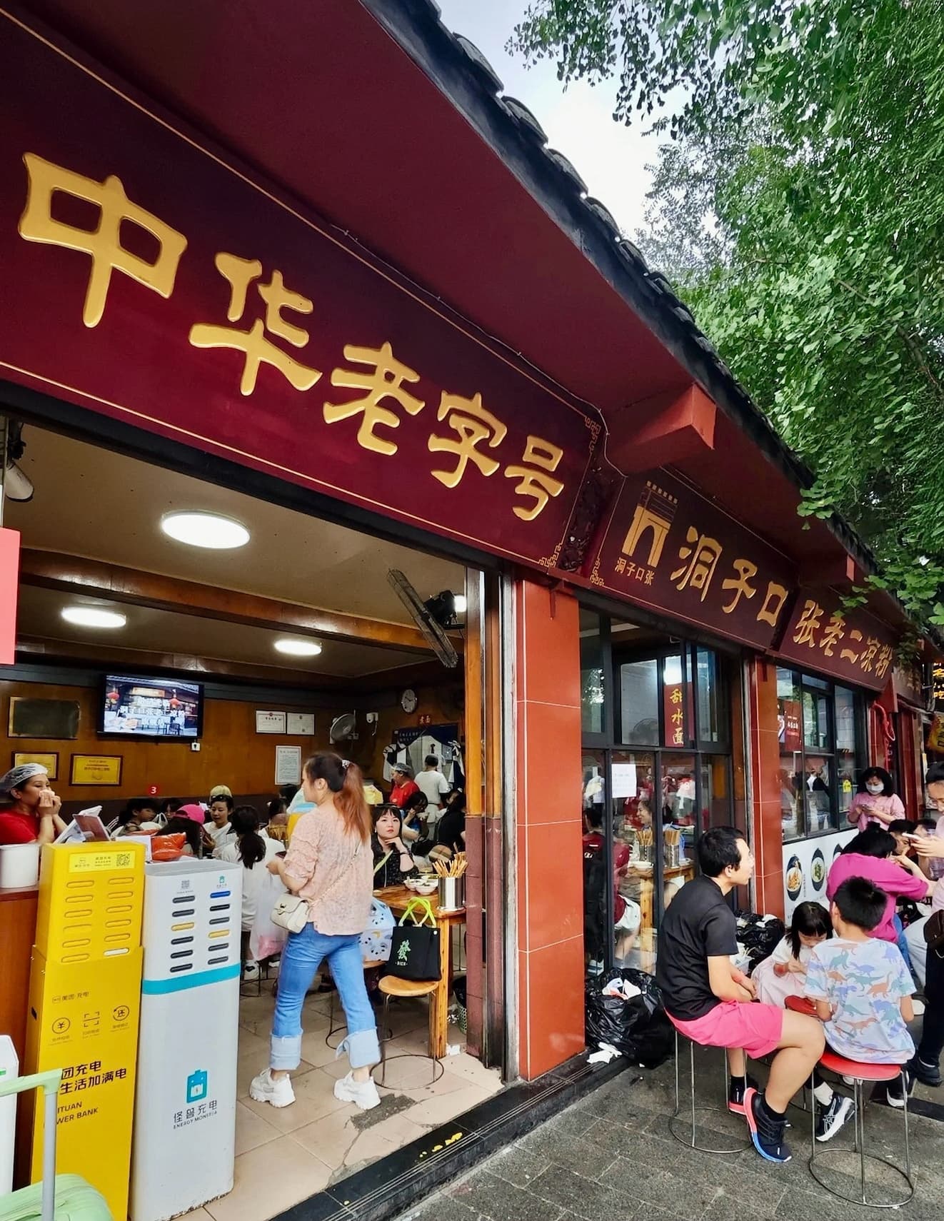 A busy street-side shop with stool seating outdoors on the sidewalk selling Chinese snacks, in Chengdu.
