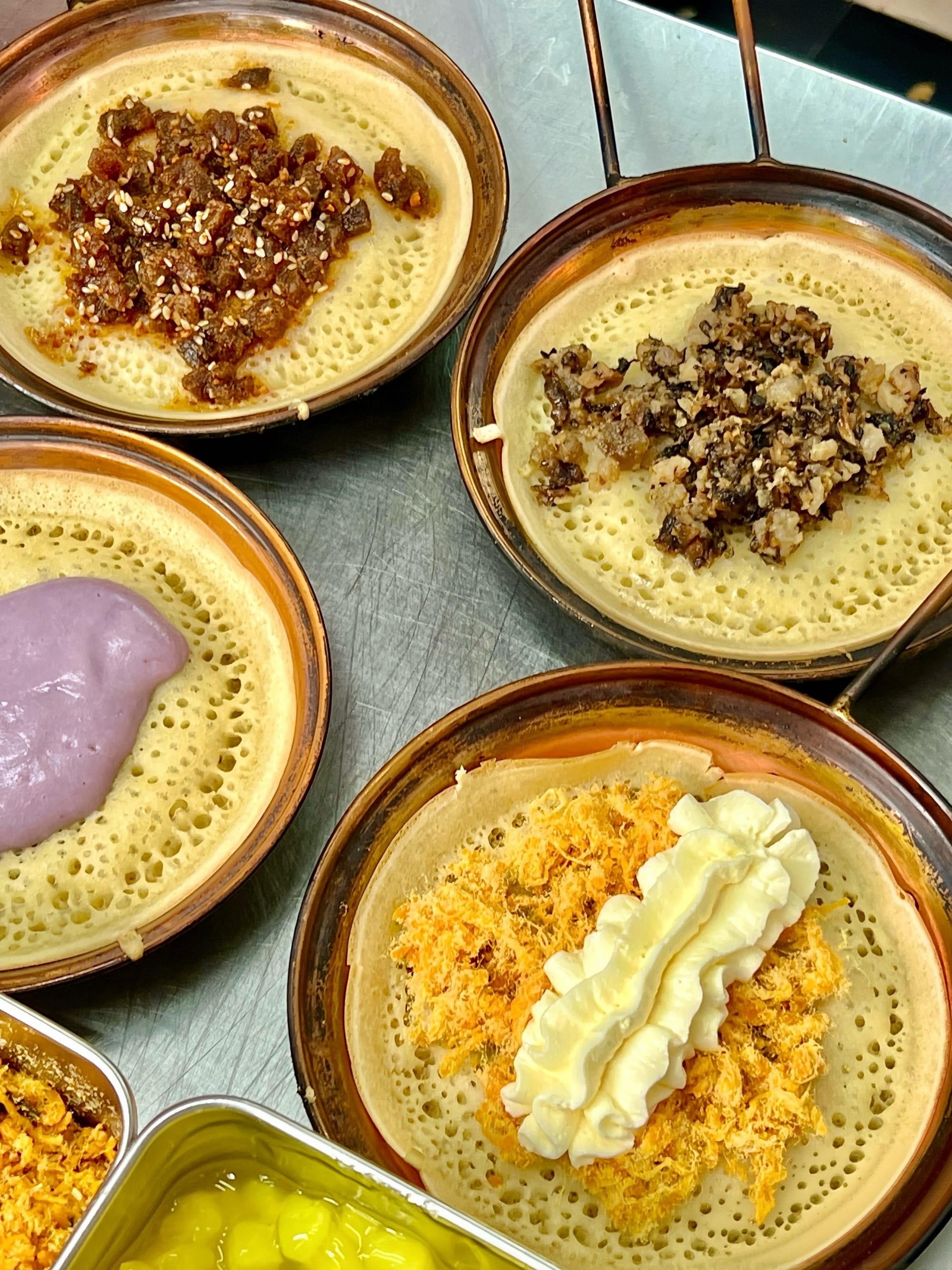 A stainless steel hot plate with small bronze frying dishes, each with a crepe and unique filling - purple paste, cream and pork floss, carmelized pork, and fermented vegetables.