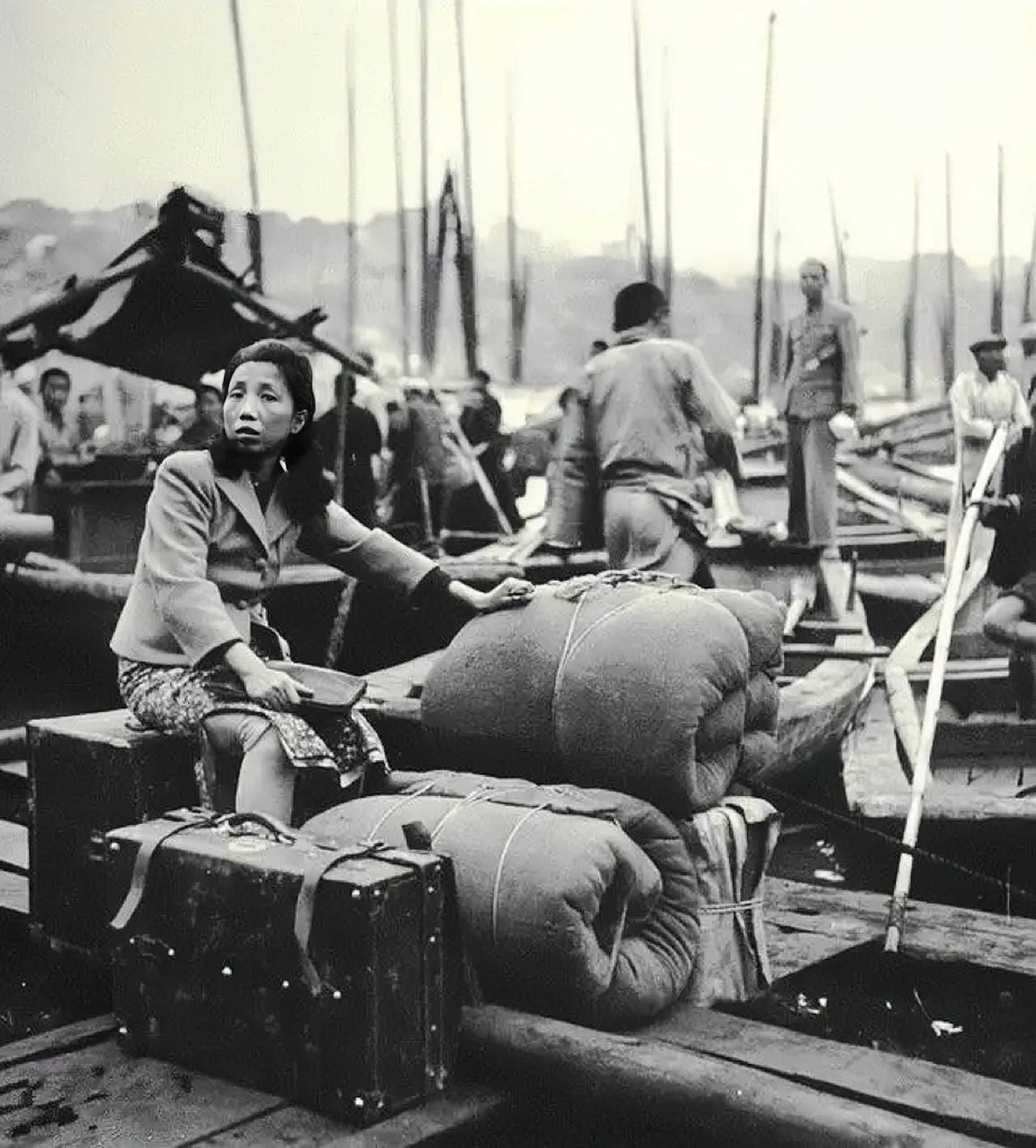 A black and white photo of a Chinese woman traveling with her old wooden suitcase and two mattress wraps rolled up, sitting on the docks of the river.