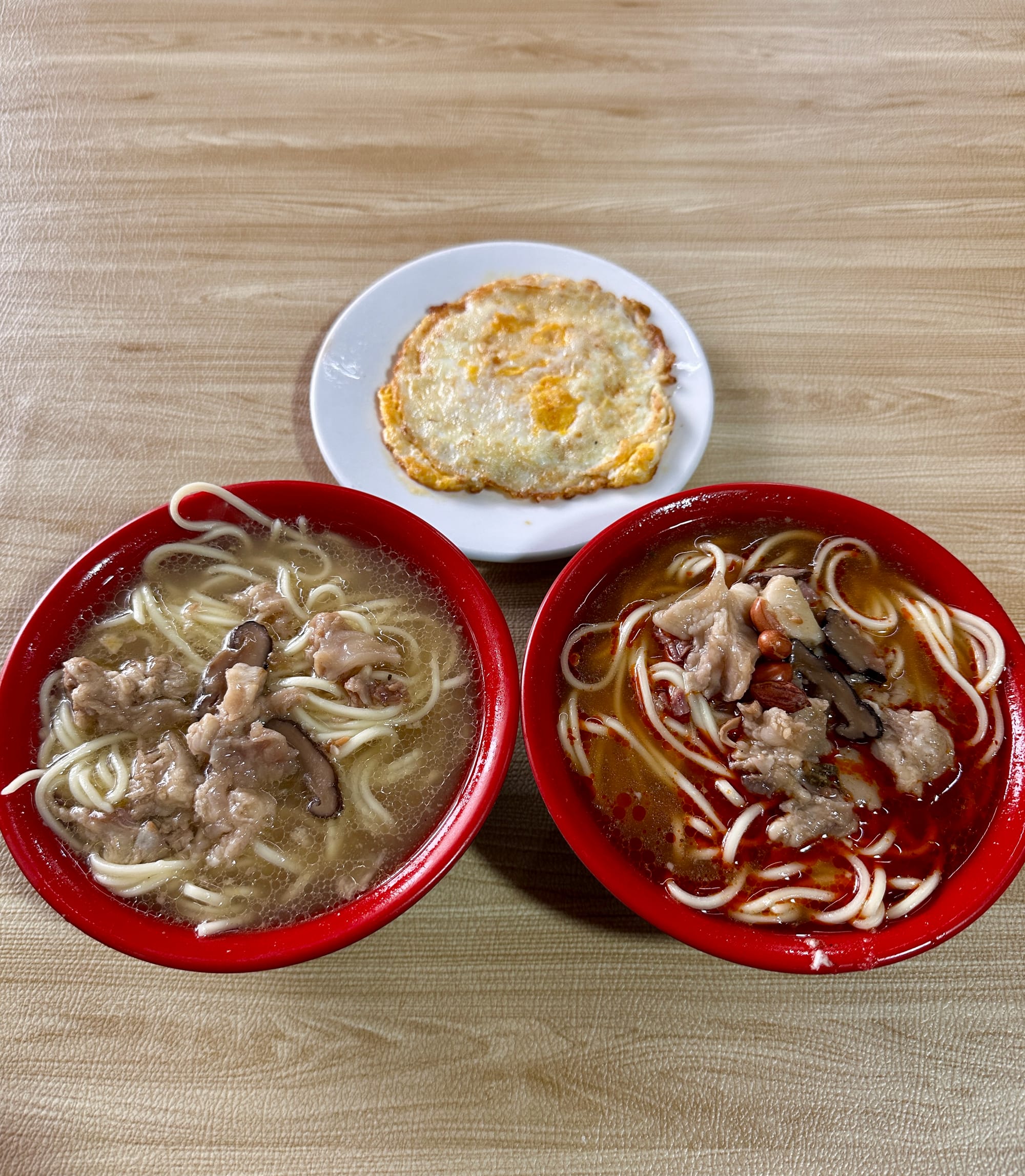 Three dishes on a table; a white dish with a fried egg, a red bowl with guaiwei noodles; and a second red bowl with haiwei noodles.