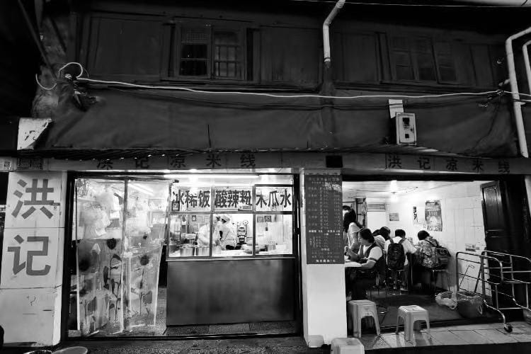 A black and white photo from the street, looking at a packed noodle shop in China.