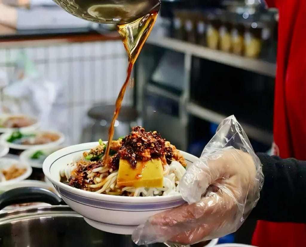 A server is ladling vinegar broth onto cold noodles with minced pork.