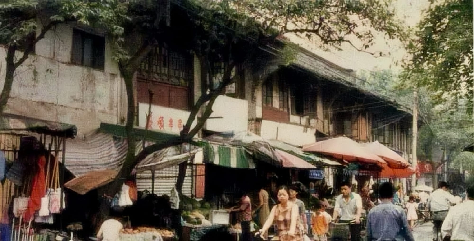 An old, weathered photo of a street in Chengdu, the ground floor of two-story buildings occupied by shops, with many people walking and biking past.