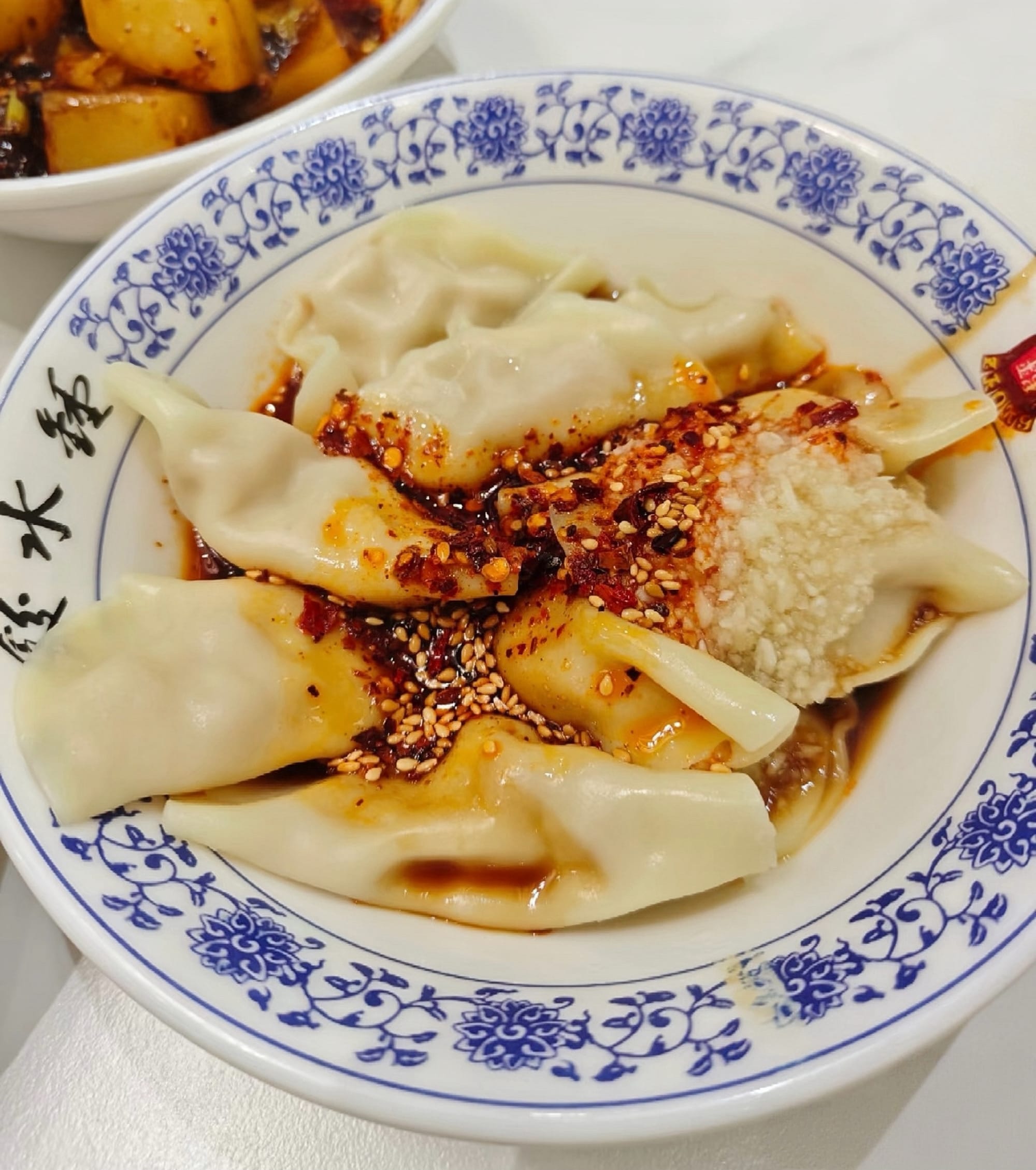 A white bowl with blue ink on the rim, contains dumplings with a red spicy sauce.