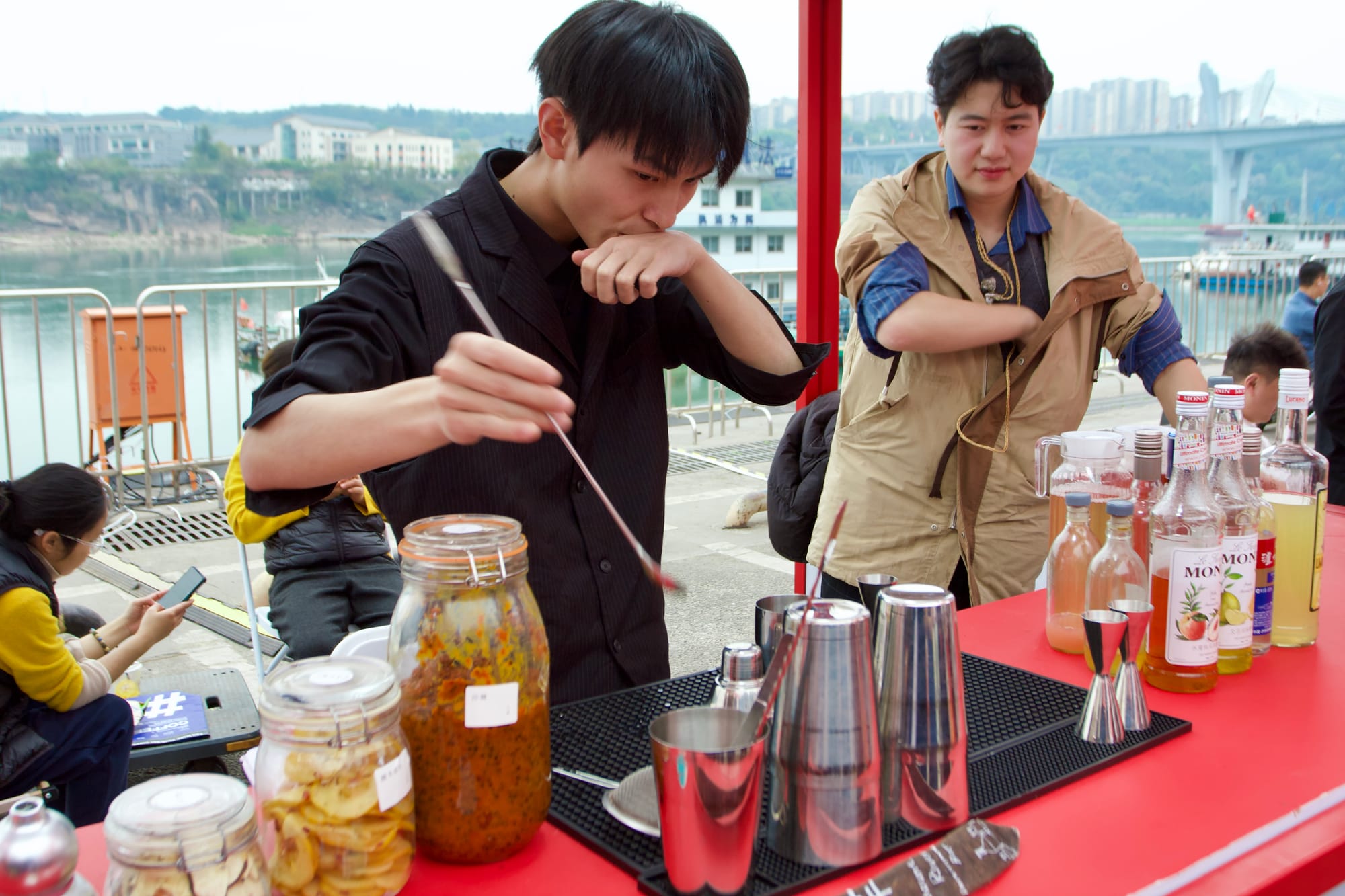 A Chinese mixologist with a long mixing utensil and an assortment of fermented dishes, cups, and alcohols on his table.
