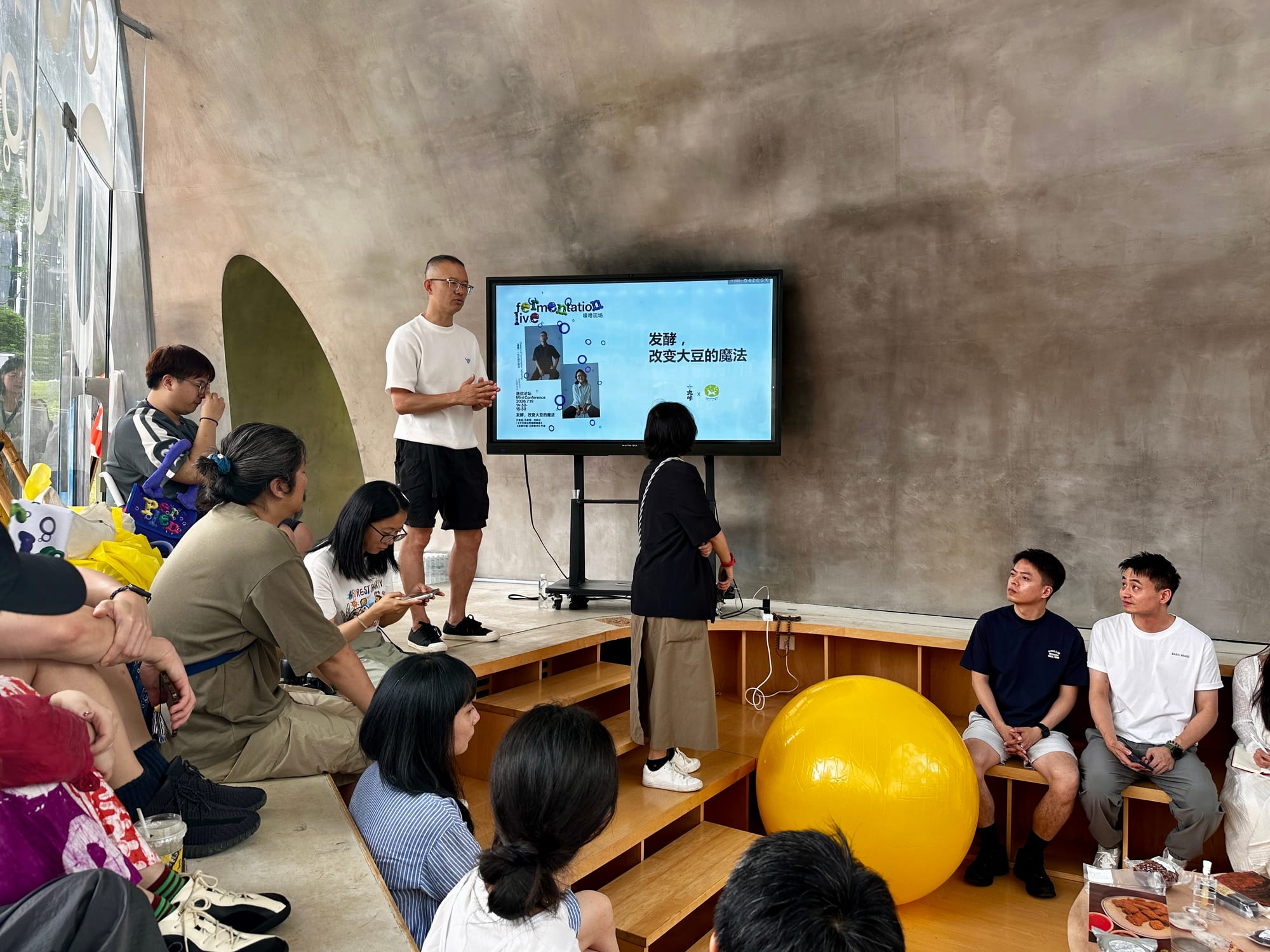 An audience sits in a semi-circle around an elevated stage watching a presenter give a talk in front of a TV