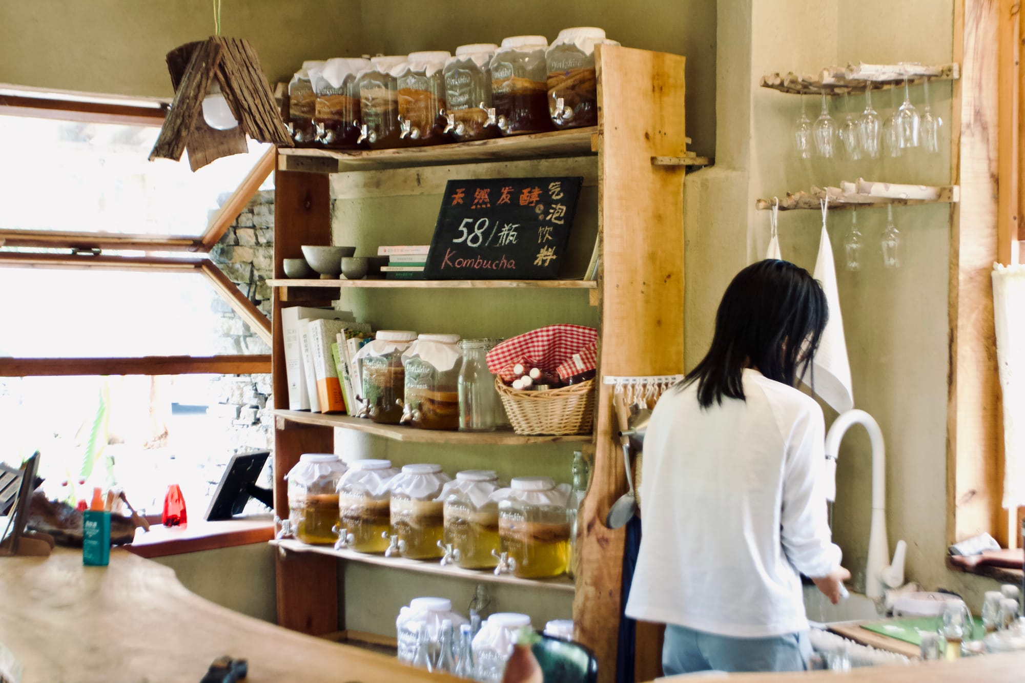 A young woman stands with her back to the camera, cleaning a glass in the sink behind a bar. On the shelves next to her are jars of kombucha, fermenting.