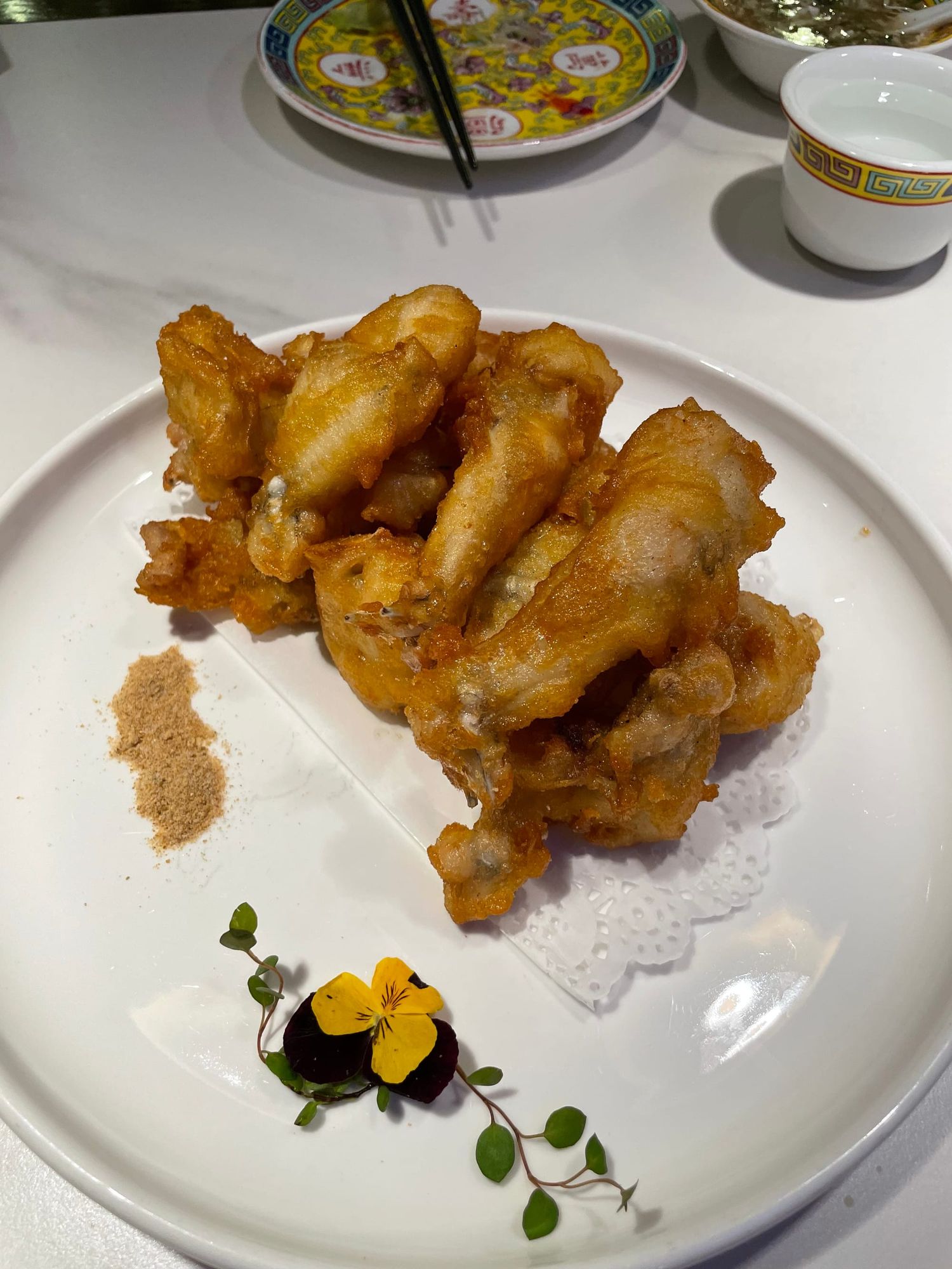 A pile of fried frog legs on a white plate next to an edible flower garnish and dry rub spice.