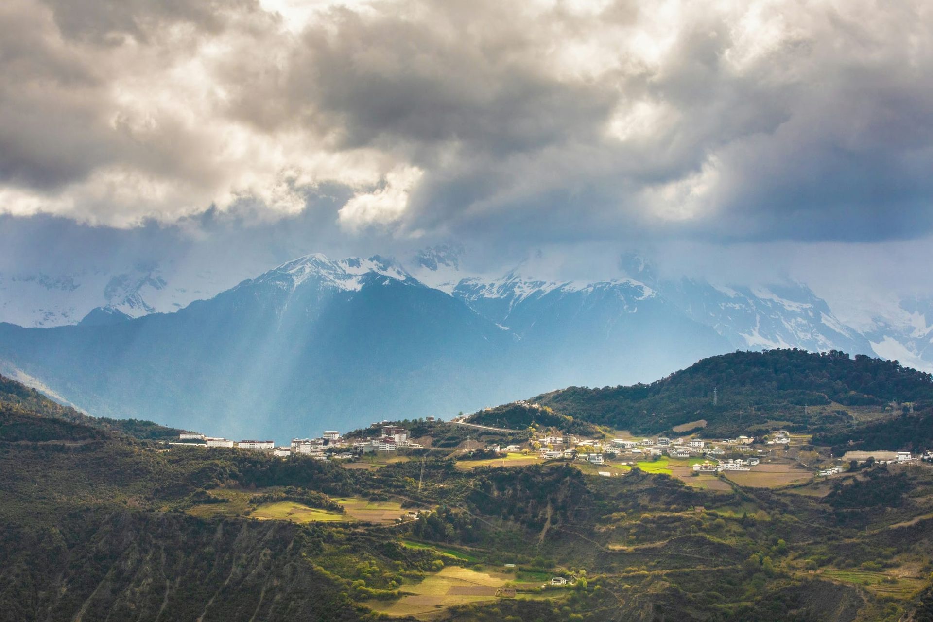A photo of Shangri-La - a green forested plateau in front of snow-capped mountains with gray clouds above with sporadic sunrays piercing through.