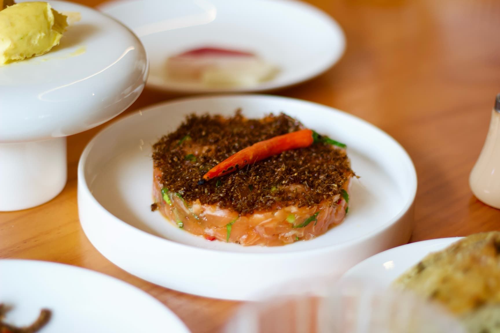 A white plate with a circular salmon tartare base topped with fermented brown ants and a pickled chili pepper