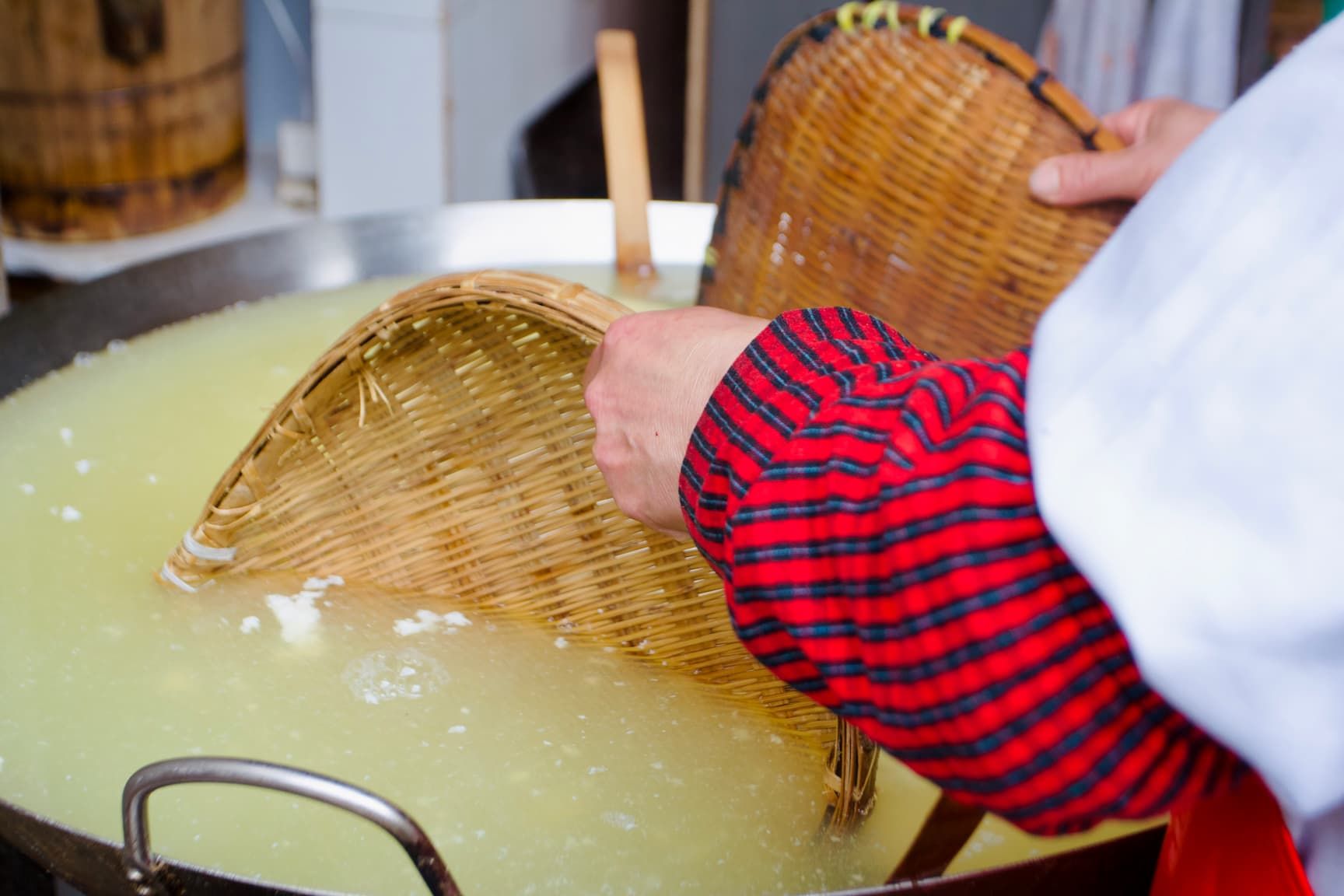 Yellow-ish clear water with small white clouds in a large stainless steel vat; a worker uses bamboo trays to wisk the water in the tofu molding process.