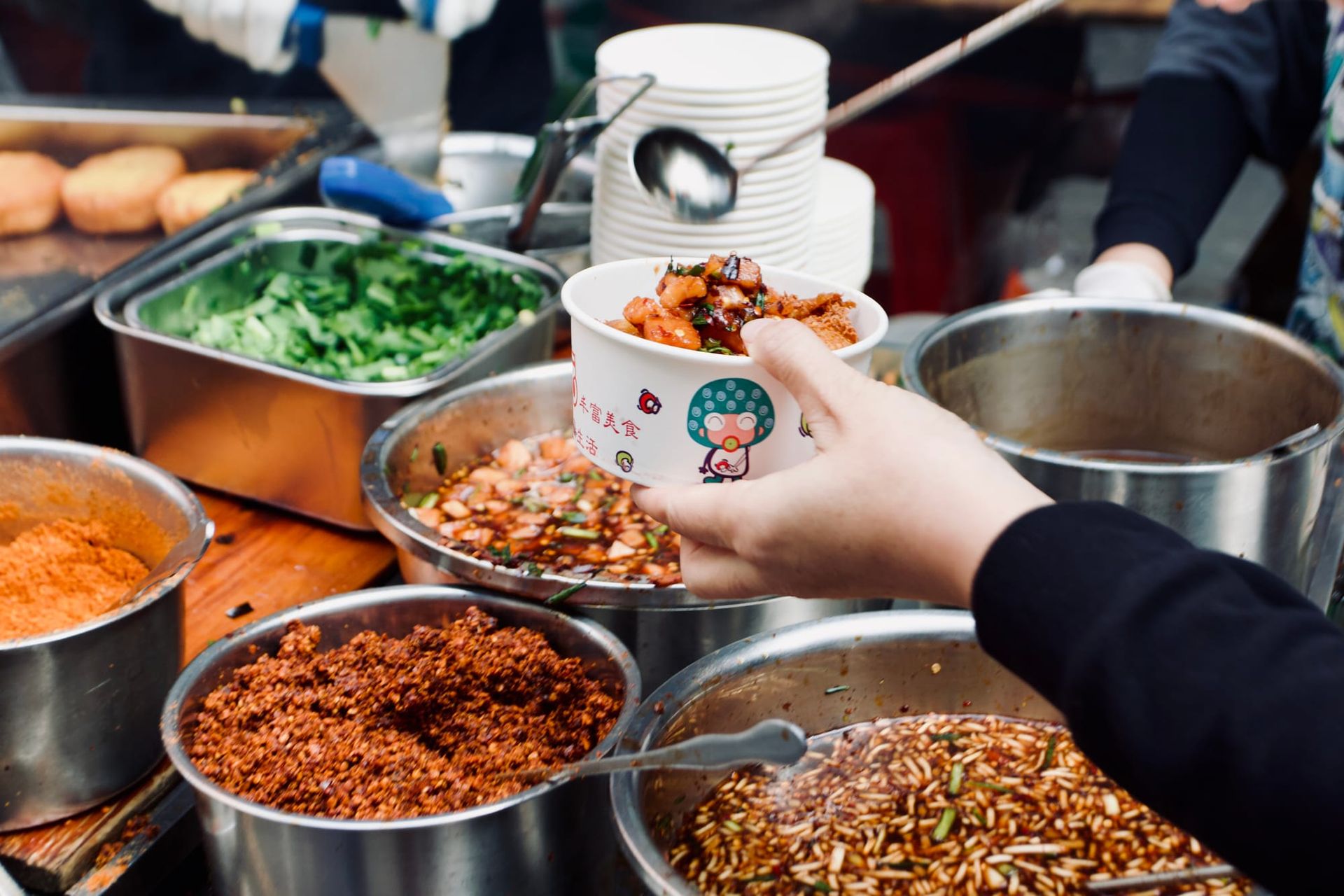 A hand holding a paper bowl with a potato dish above many stainless steel pots with condiments.