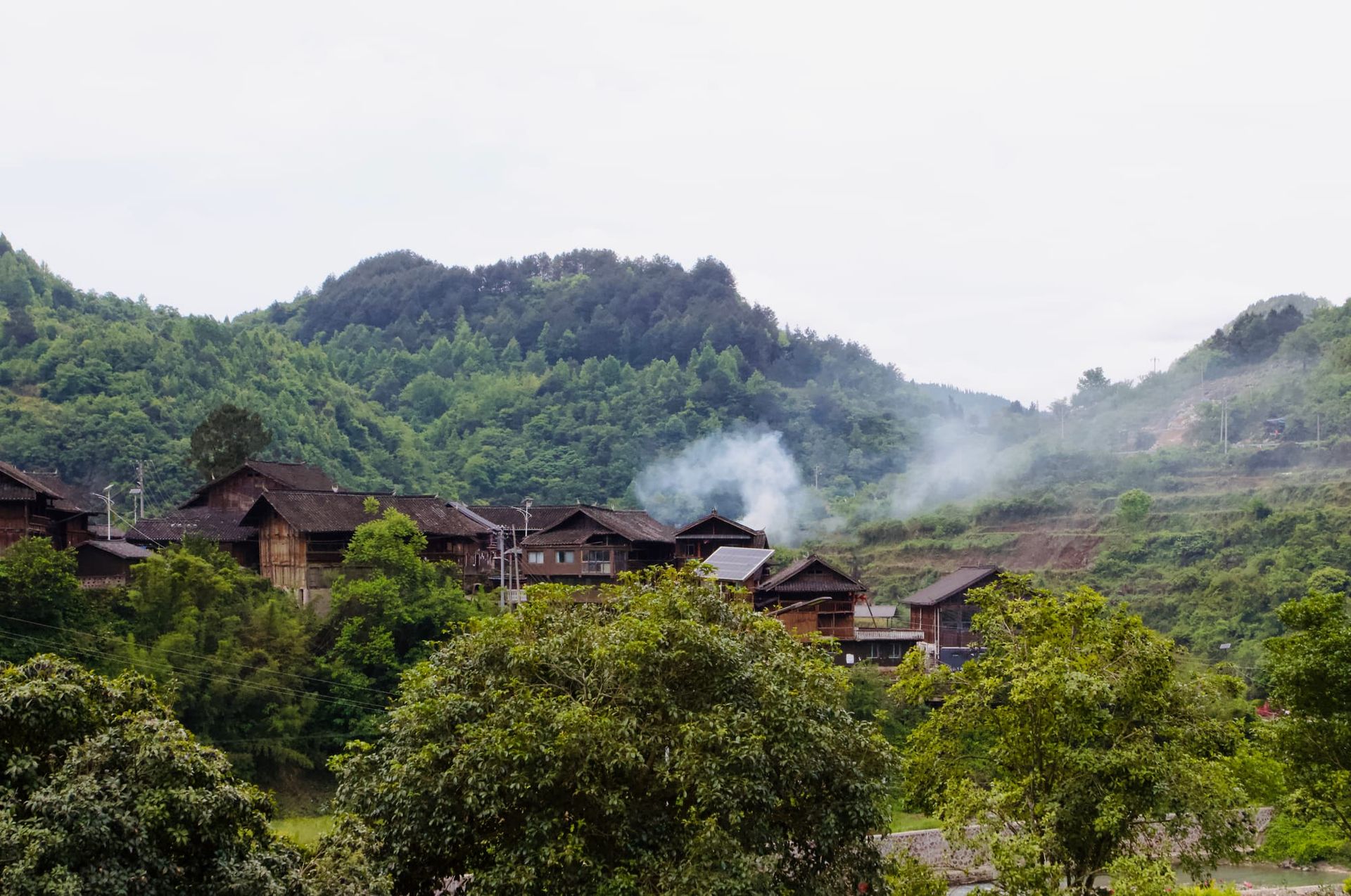 Green mountains with brown wooden houses nestled in the middle, as smoke rises from cooking fires.